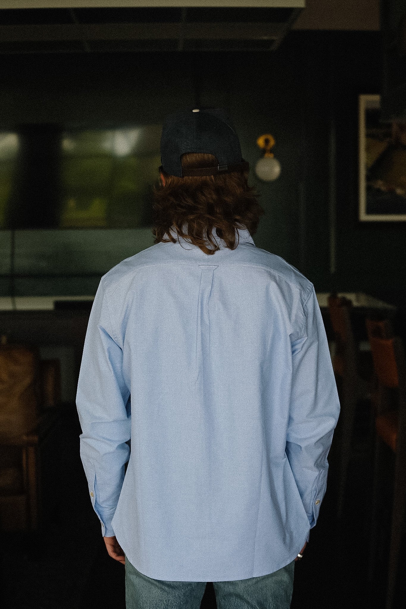 Person wearing a light blue shirt and dark cap in a dimly lit room.