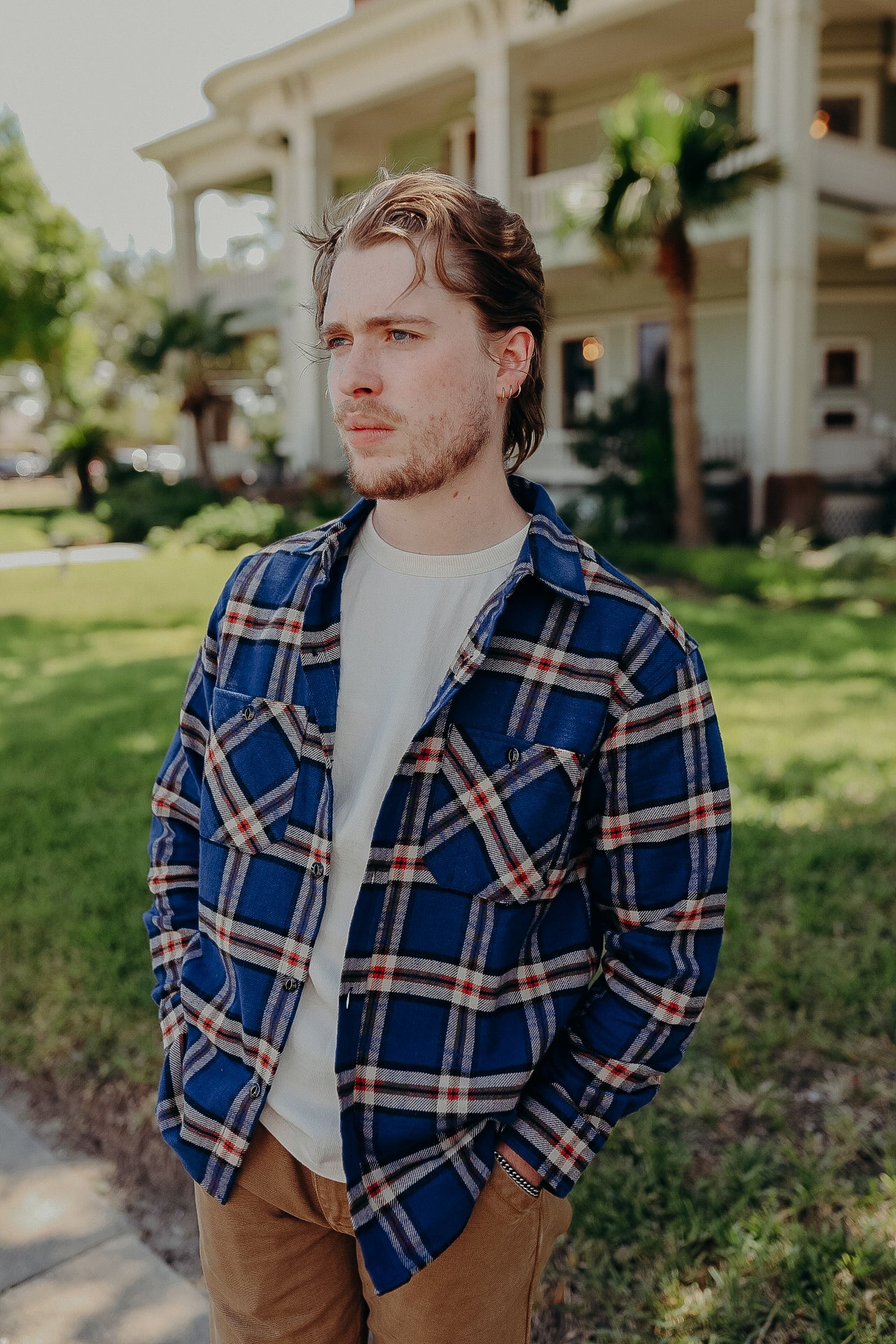 Man wearing blue plaid flannel shirt outdoors in front of a building with palm trees, casual iron provision style