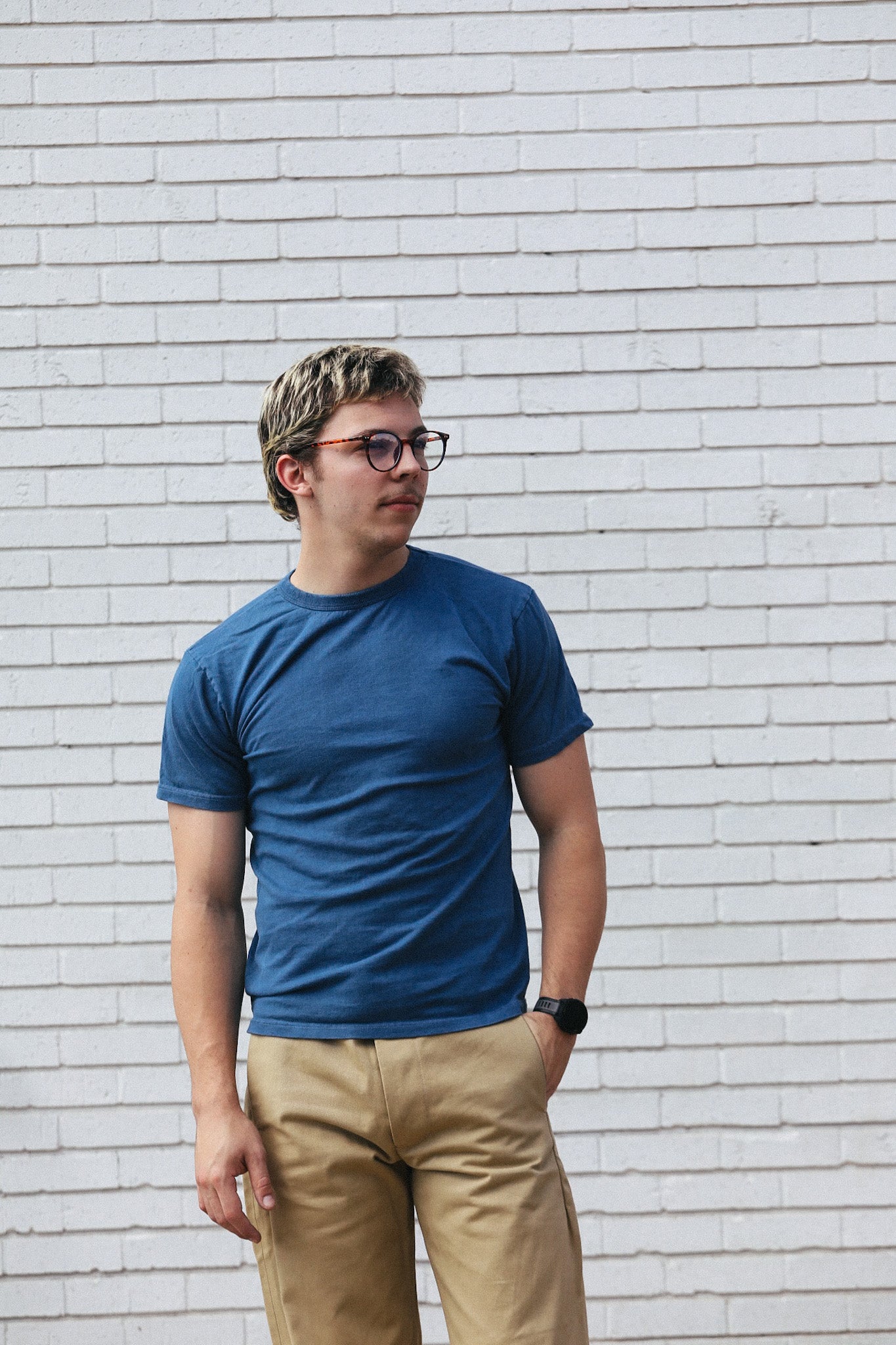 Young man wearing a blue t-shirt and beige pants standing against a white brick wall