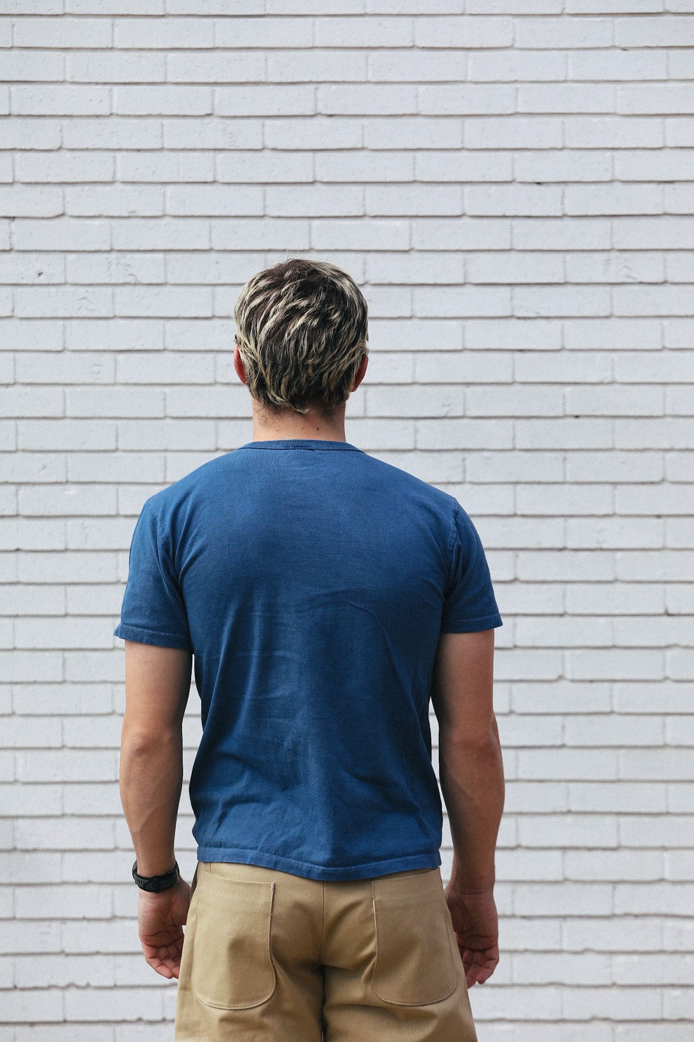 Man with short blond hair wearing blue t-shirt and beige pants standing against white brick wall