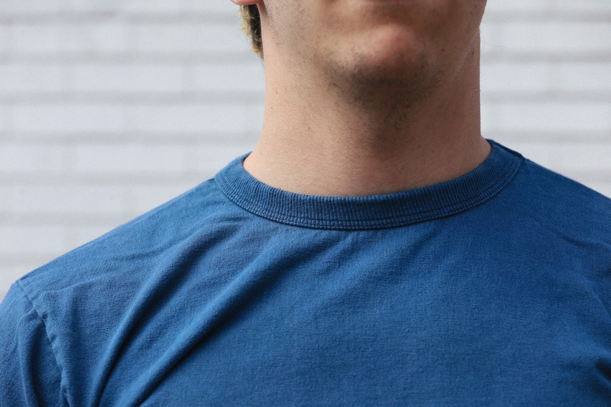 Close-up of a man wearing a blue crewneck t-shirt in front of a light gray brick wall