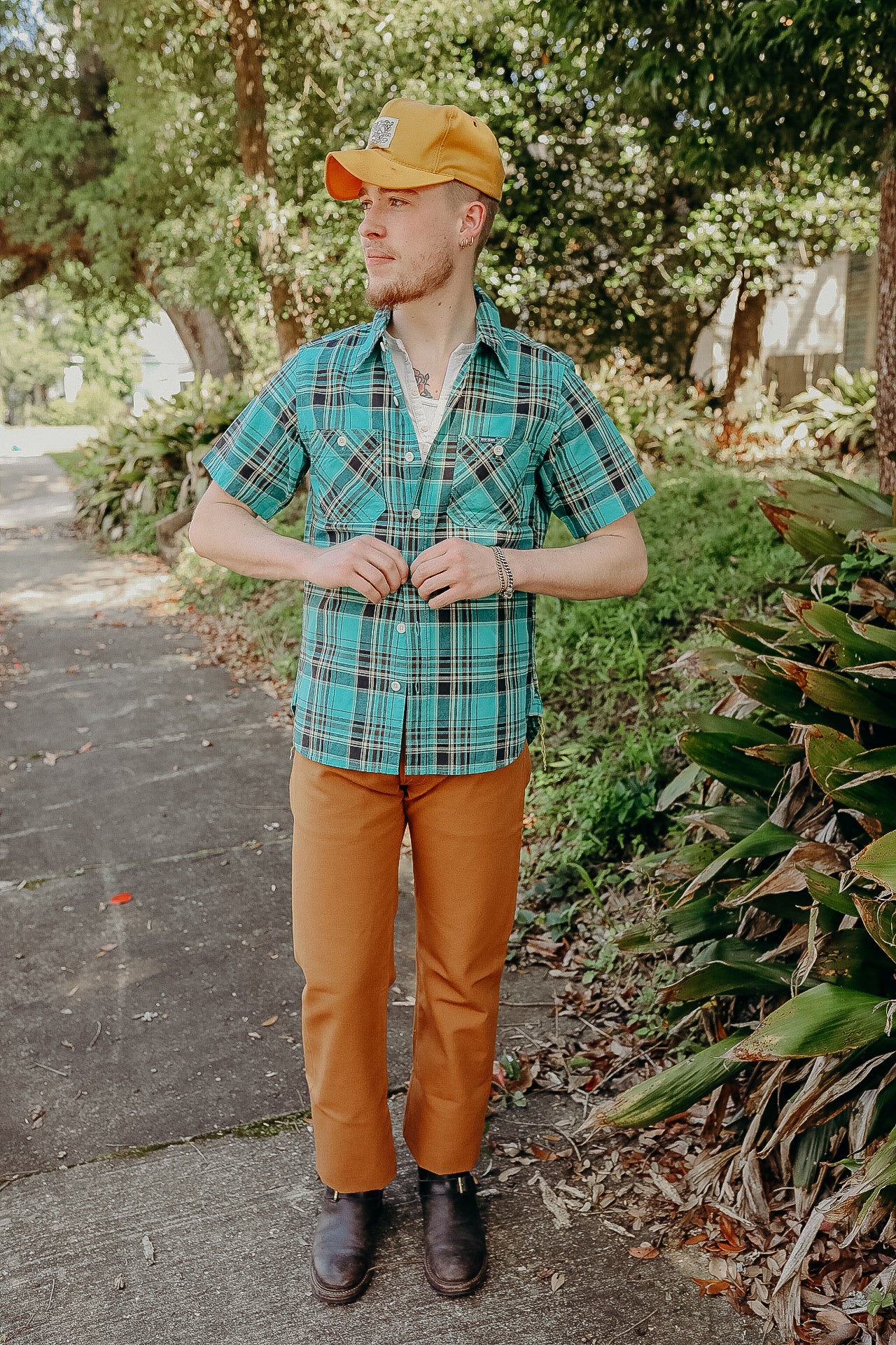 Man wearing turquoise plaid short sleeve shirt, mustard pants, and mustard cap in leafy outdoor setting