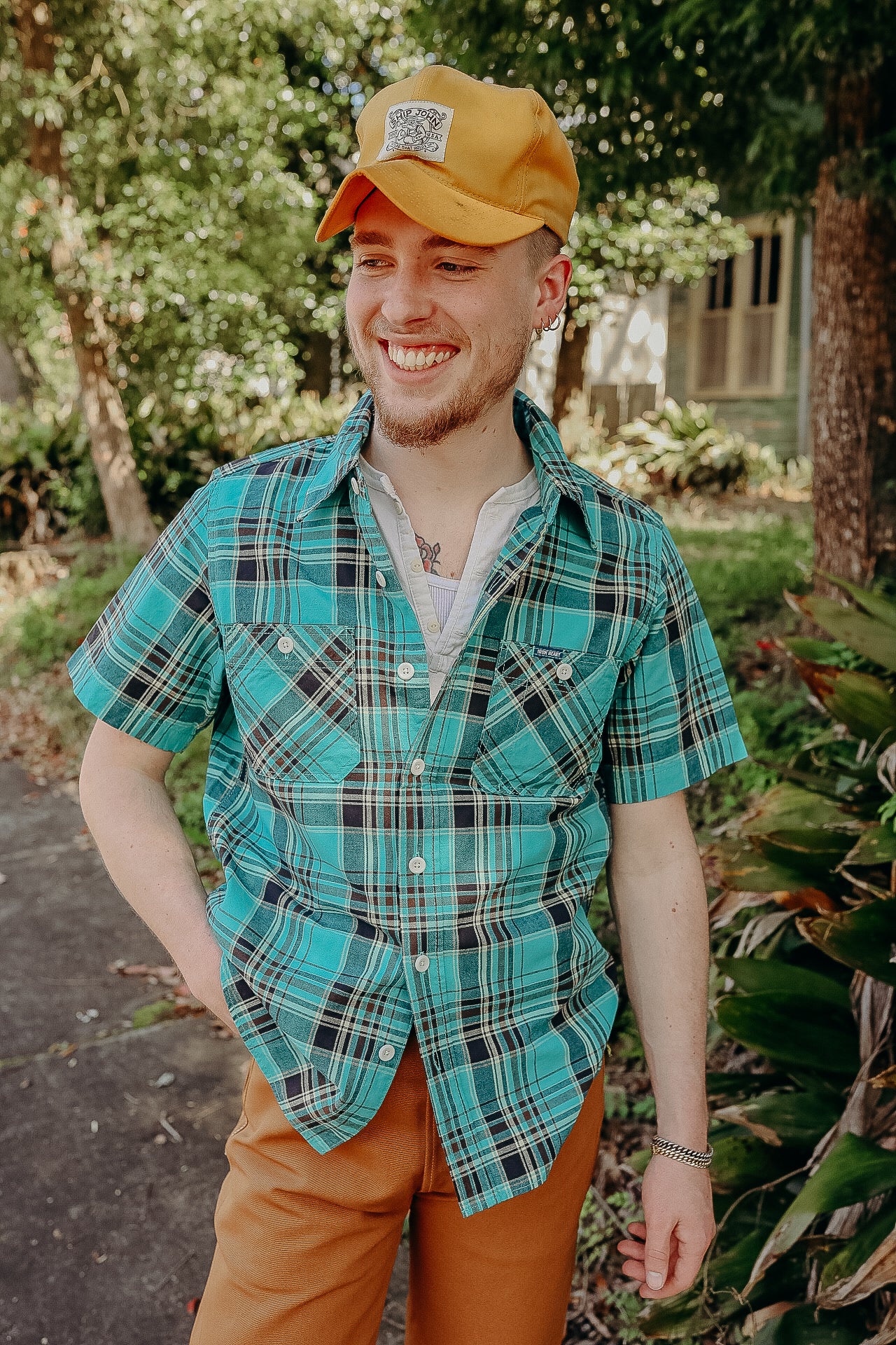 Smiling young man in a turquoise plaid short-sleeve shirt, mustard cap, and brown pants outdoors in a sunny garden setting