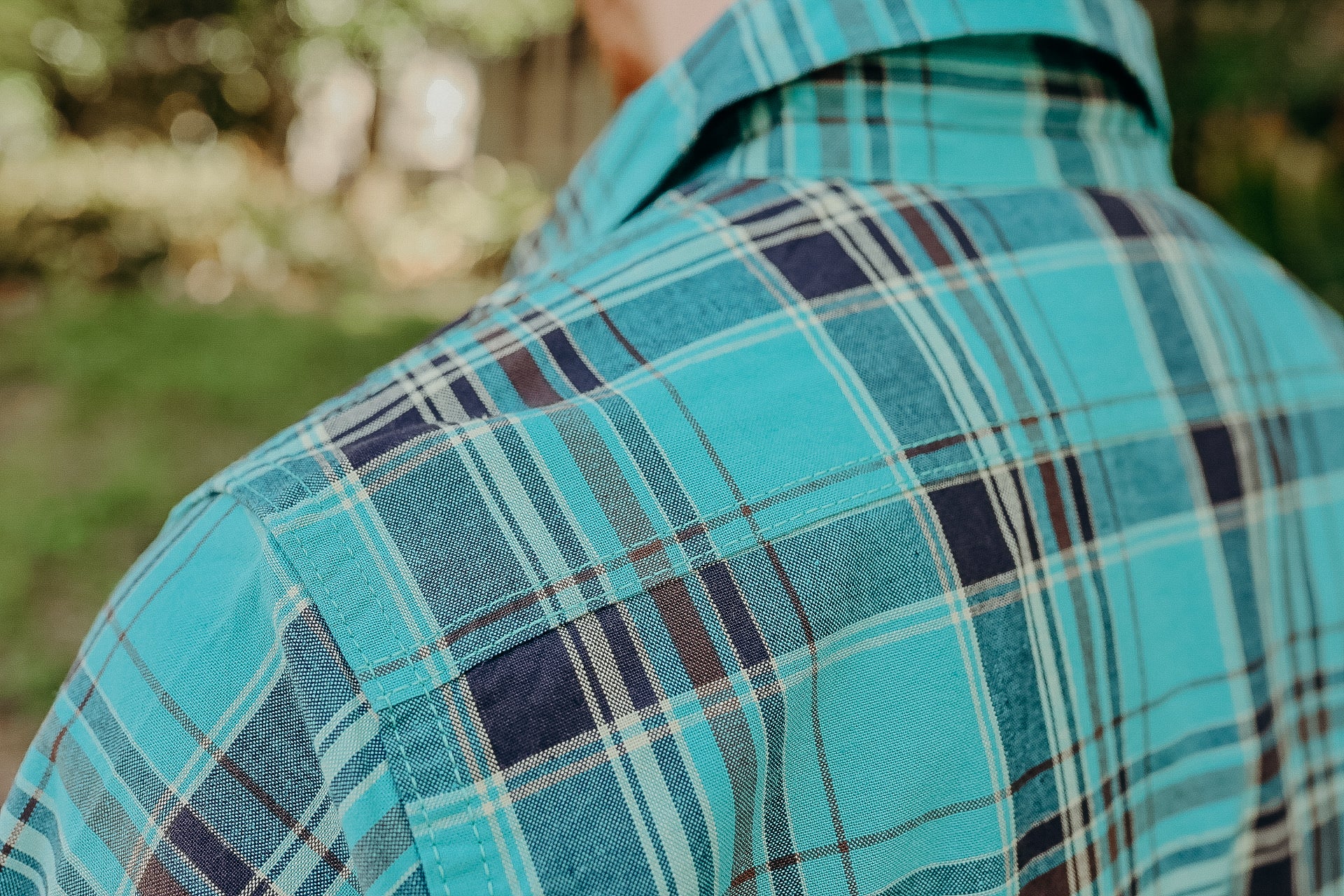 Close-up of a turquoise and navy blue plaid shirt shoulder with outdoor blurred background