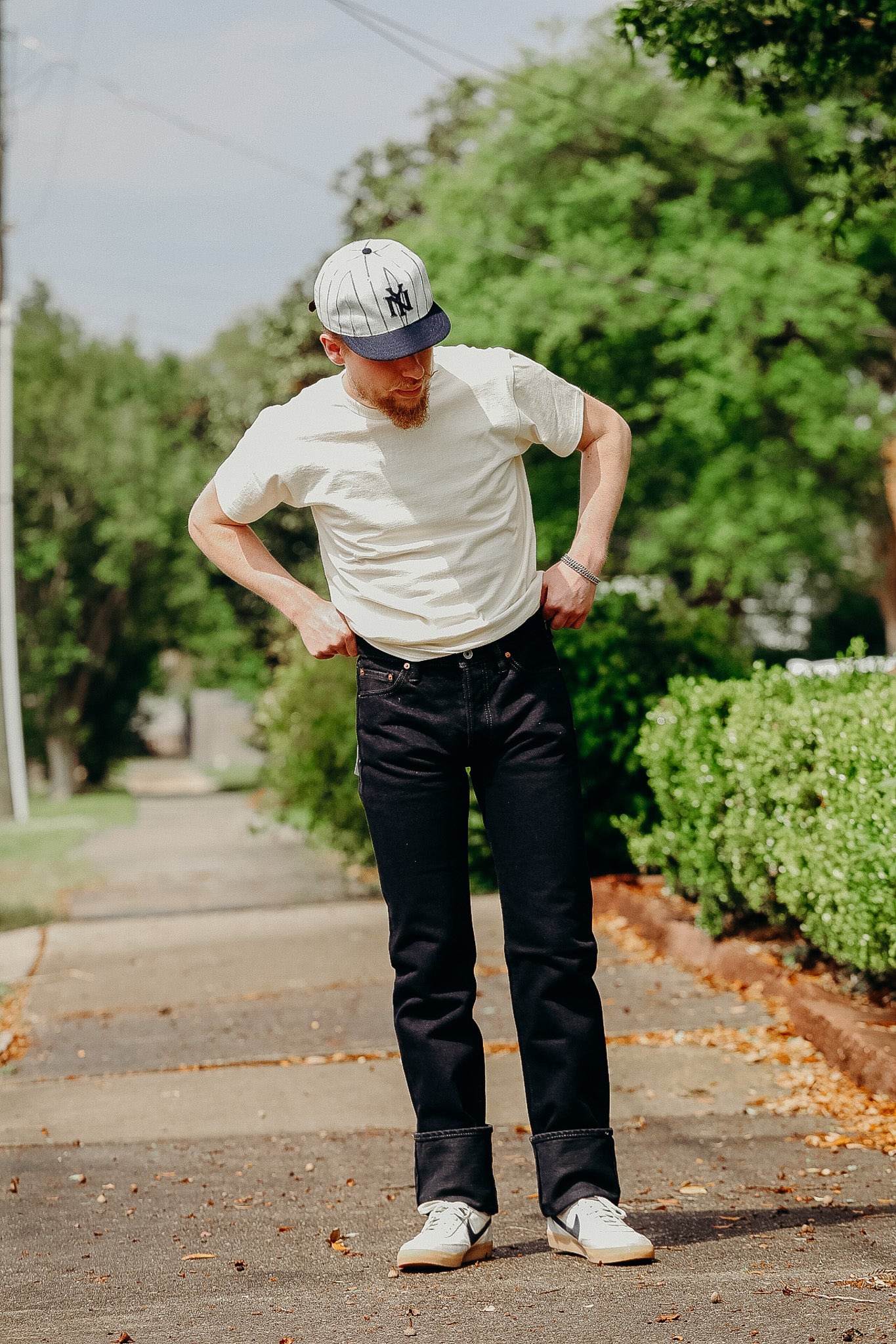 Man wearing black jeans, white t-shirt, and baseball cap standing on a sidewalk with green trees