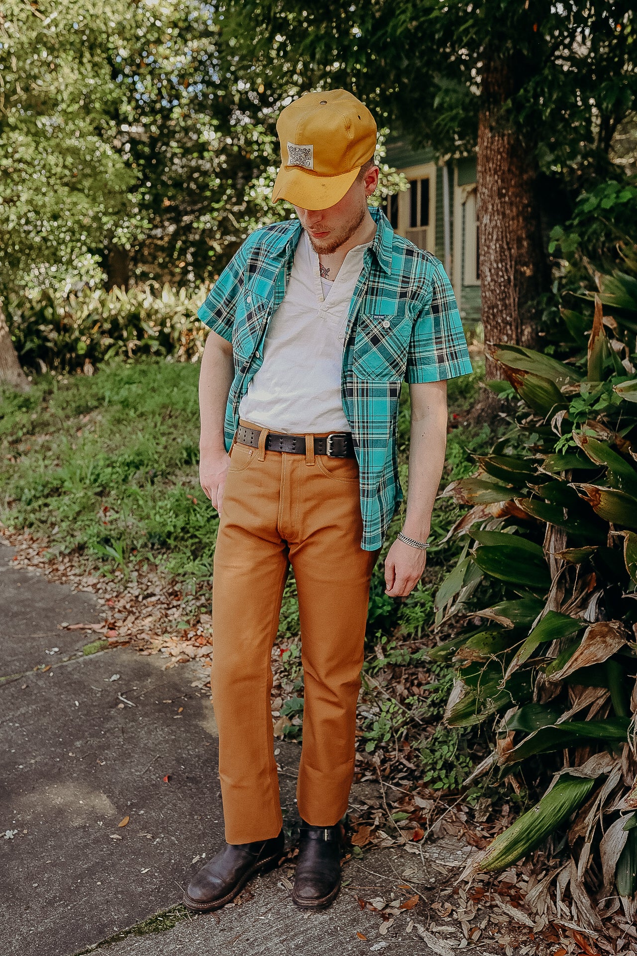Man wearing mustard yellow pants, blue plaid shirt, white t-shirt, yellow cap, and black boots outdoors