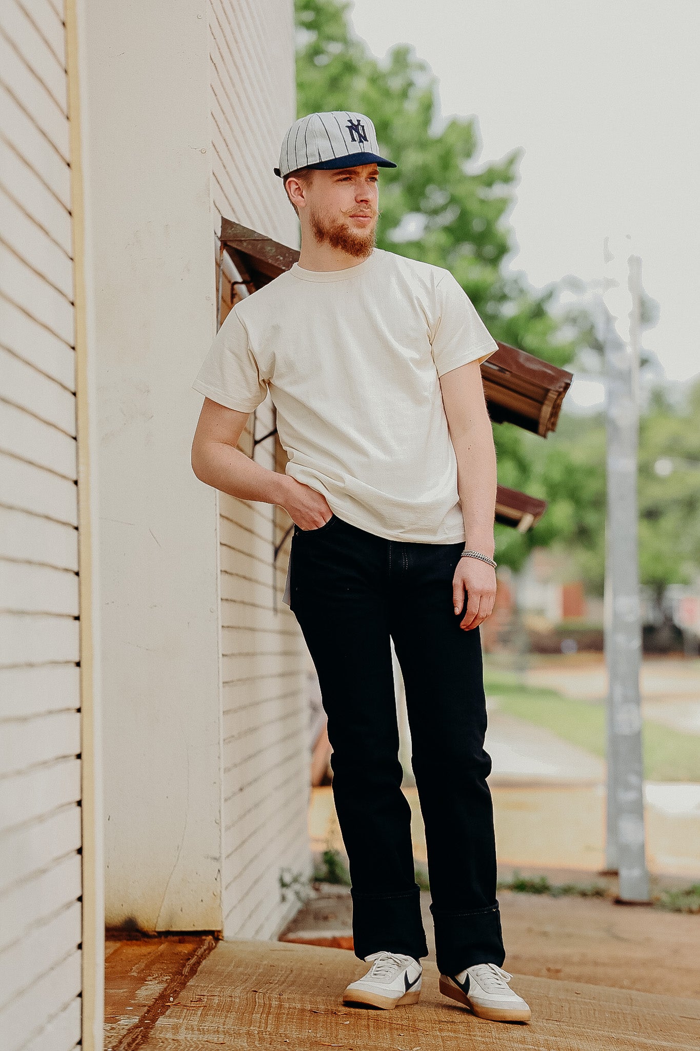 Man wearing beige t-shirt, black pants, Nike sneakers, and NY cap leaning against white building outdoors