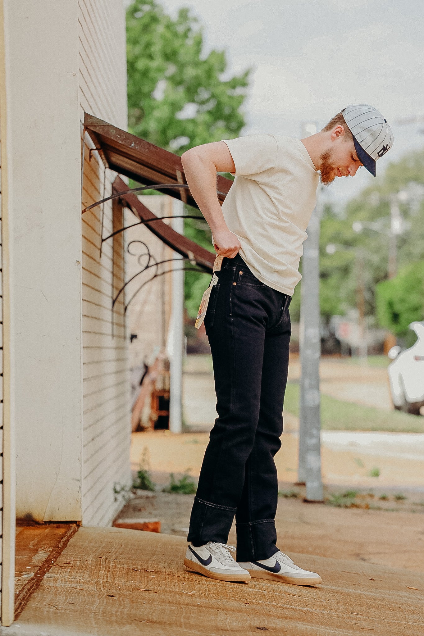 Man wearing white t-shirt, black jeans, and sneakers, adjusting jeans tags outdoors near brick wall