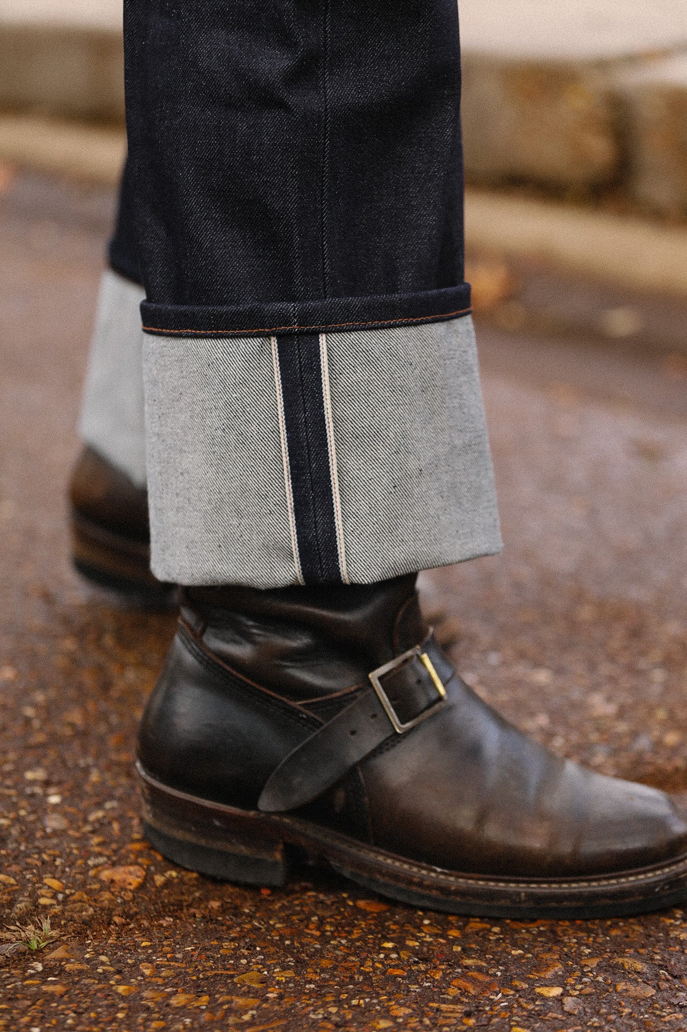 Close-up of raw selvedge denim cuff paired with dark brown leather engineer boots on textured ground