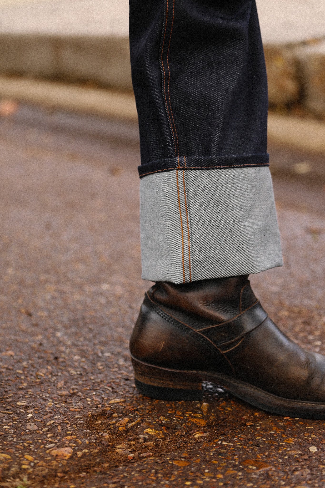 Cuffed dark raw denim jeans paired with brown leather ankle boots on gravel surface