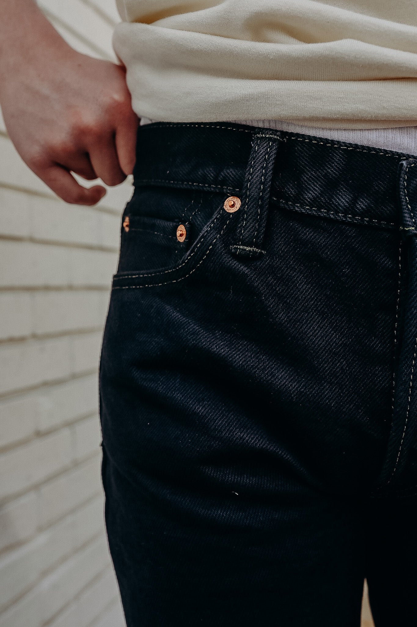 Close-up of person wearing dark denim jeans with copper rivets and white shirt against a brick wall