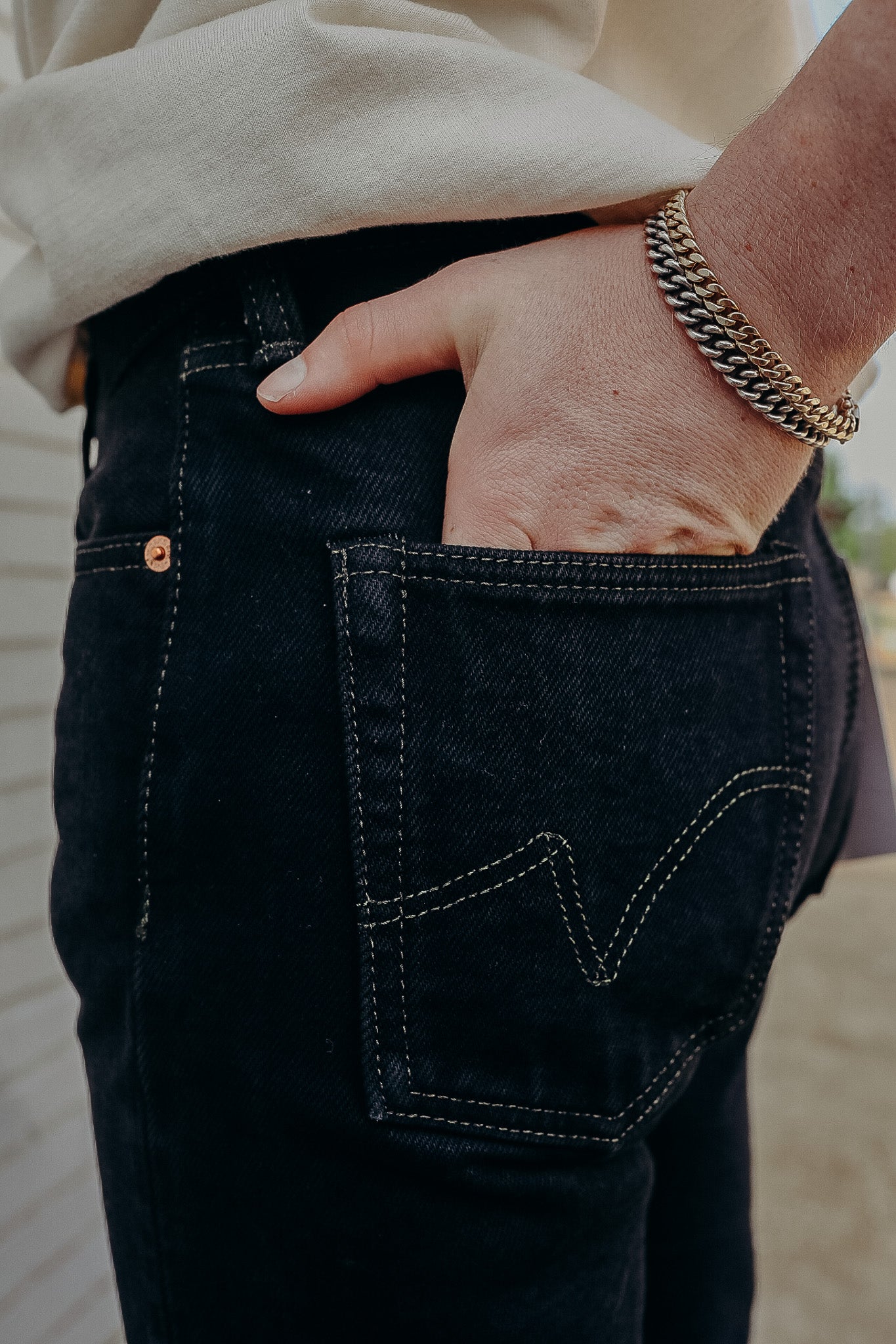 Close-up of hand wearing gold and silver chain bracelets resting in back pocket of black denim jeans with white stitching