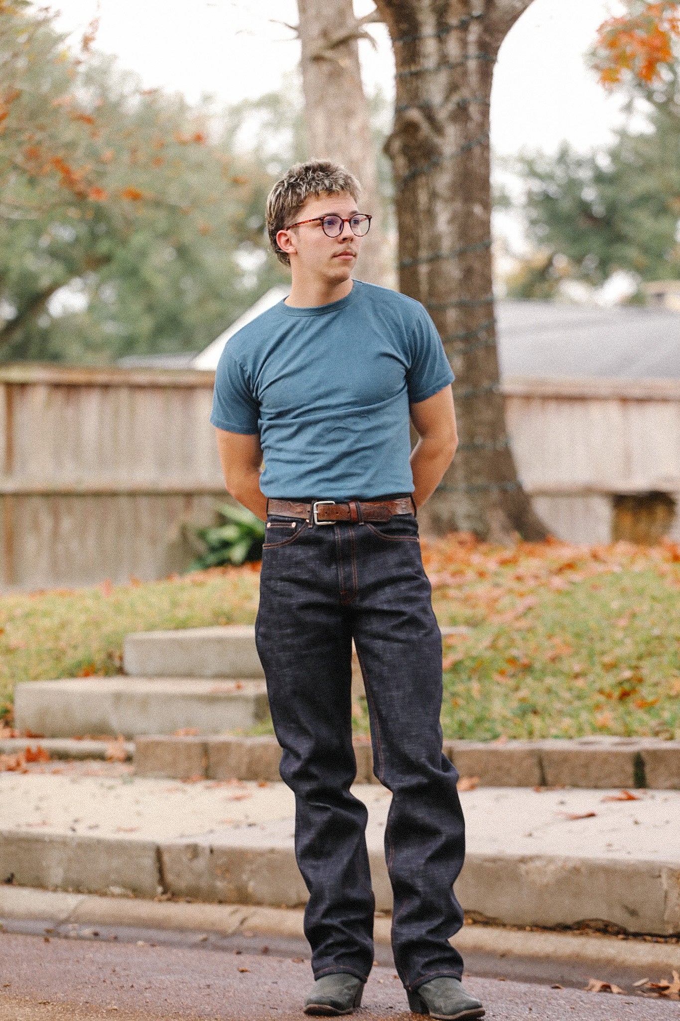 Man wearing blue t-shirt and dark indigo wide straight jeans standing outdoors with trees and fallen leaves
