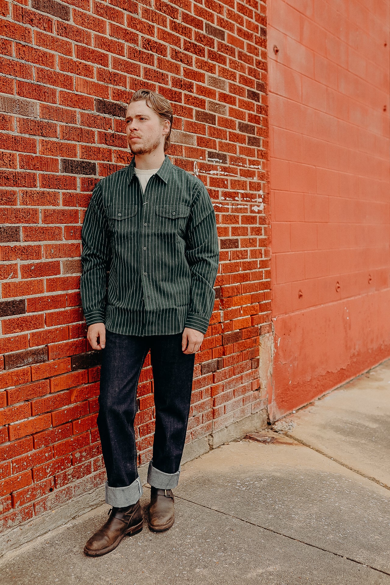 Man wearing green striped work shirt, dark rolled-up jeans, and brown boots standing against red brick wall