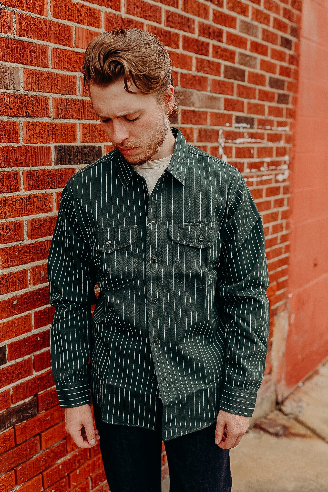 Man wearing green pinstripe button-up shirt standing against red brick wall at Iron Shop Provisions