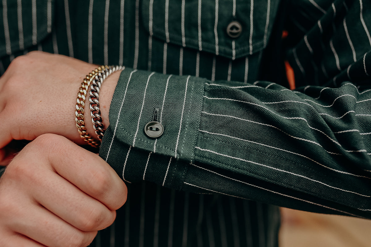 Close-up of a wrist with gold and silver chain bracelets wearing a green striped shirt from Iron Shop Provisions