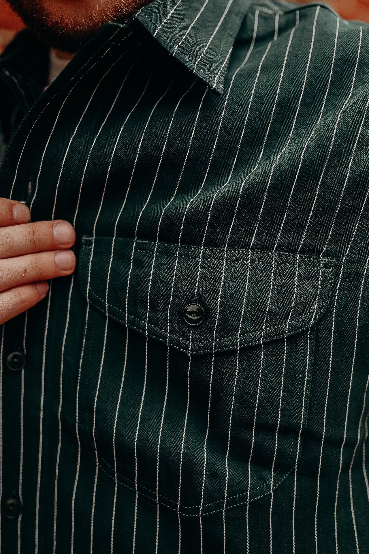 Close-up of green pinstripe shirt pocket with black button and hand adjusting shirt near collar
