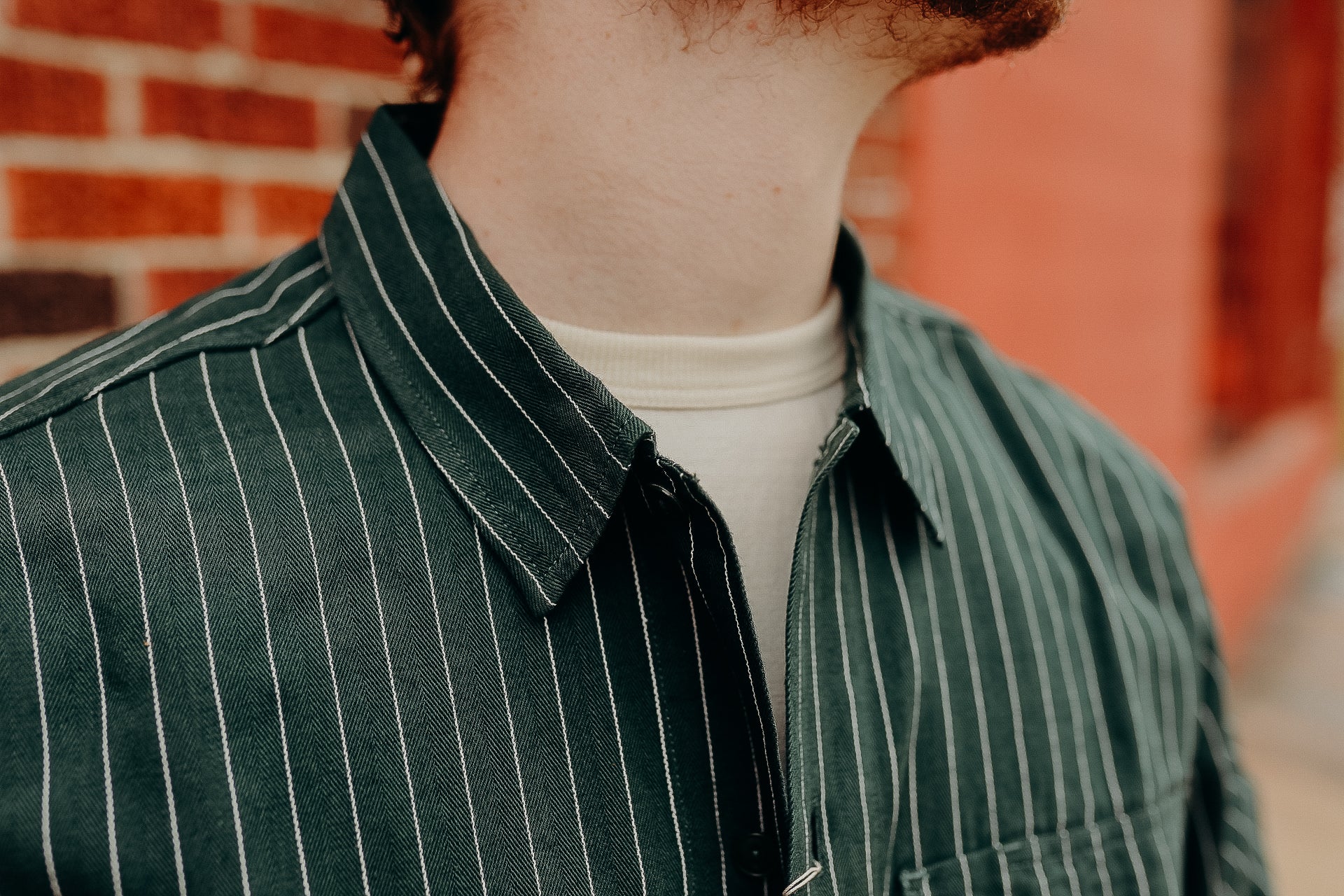 Close-up of person wearing green striped Iron Shop Provisions shirt over white undershirt by brick wall