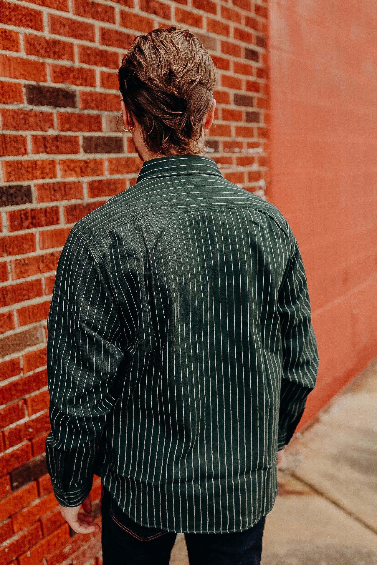Man wearing dark green pinstripe shirt standing against red brick and painted wall outdoors
