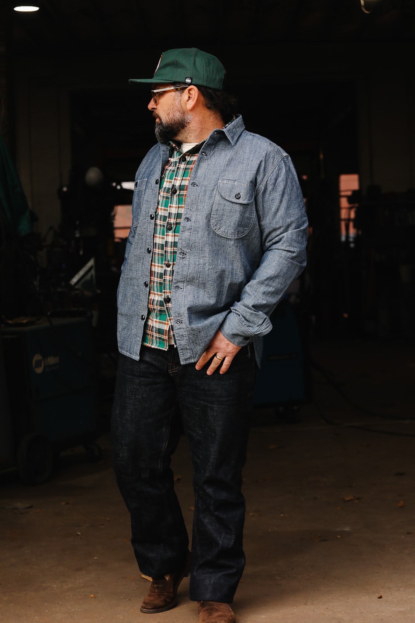 Man wearing a green cap, glasses, blue denim jacket, green plaid shirt, dark jeans, and brown boots in a workshop