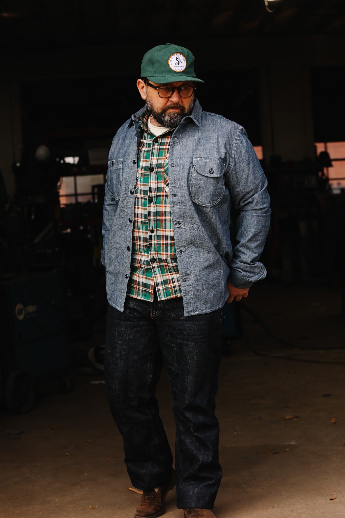 Man wearing green cap, glasses, plaid shirt, denim jacket and dark jeans in workshop setting