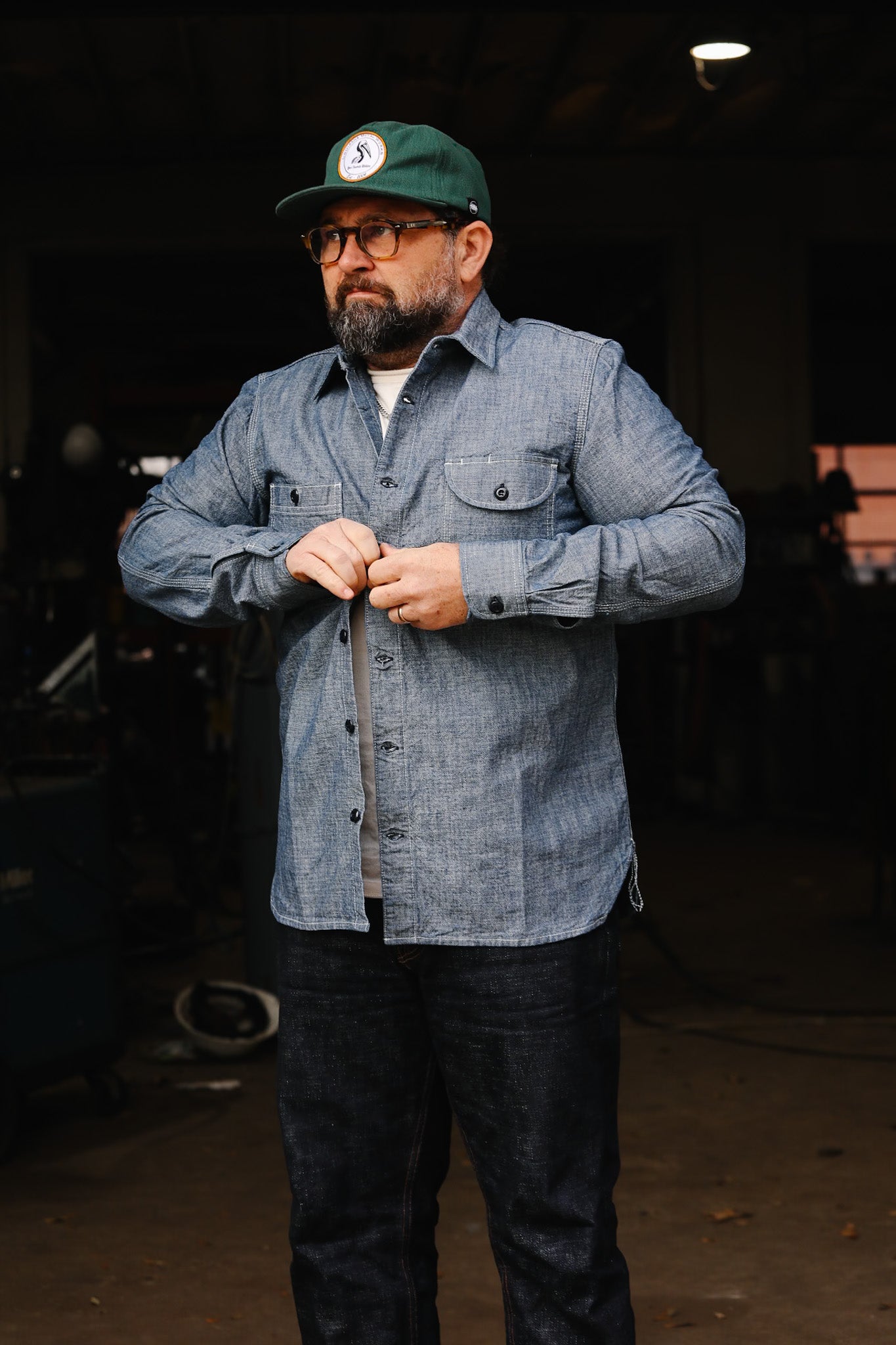 Man wearing green cap and blue chambray work shirt buttoning up in industrial workshop
