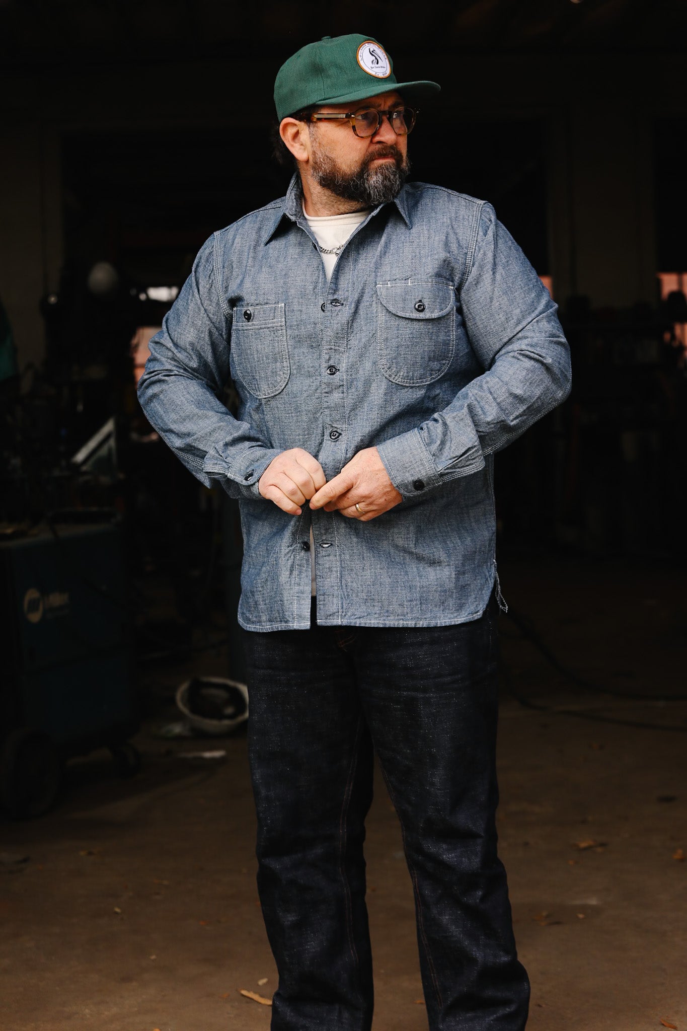 Man wearing denim work shirt, dark jeans, green cap, and glasses in workshop setting