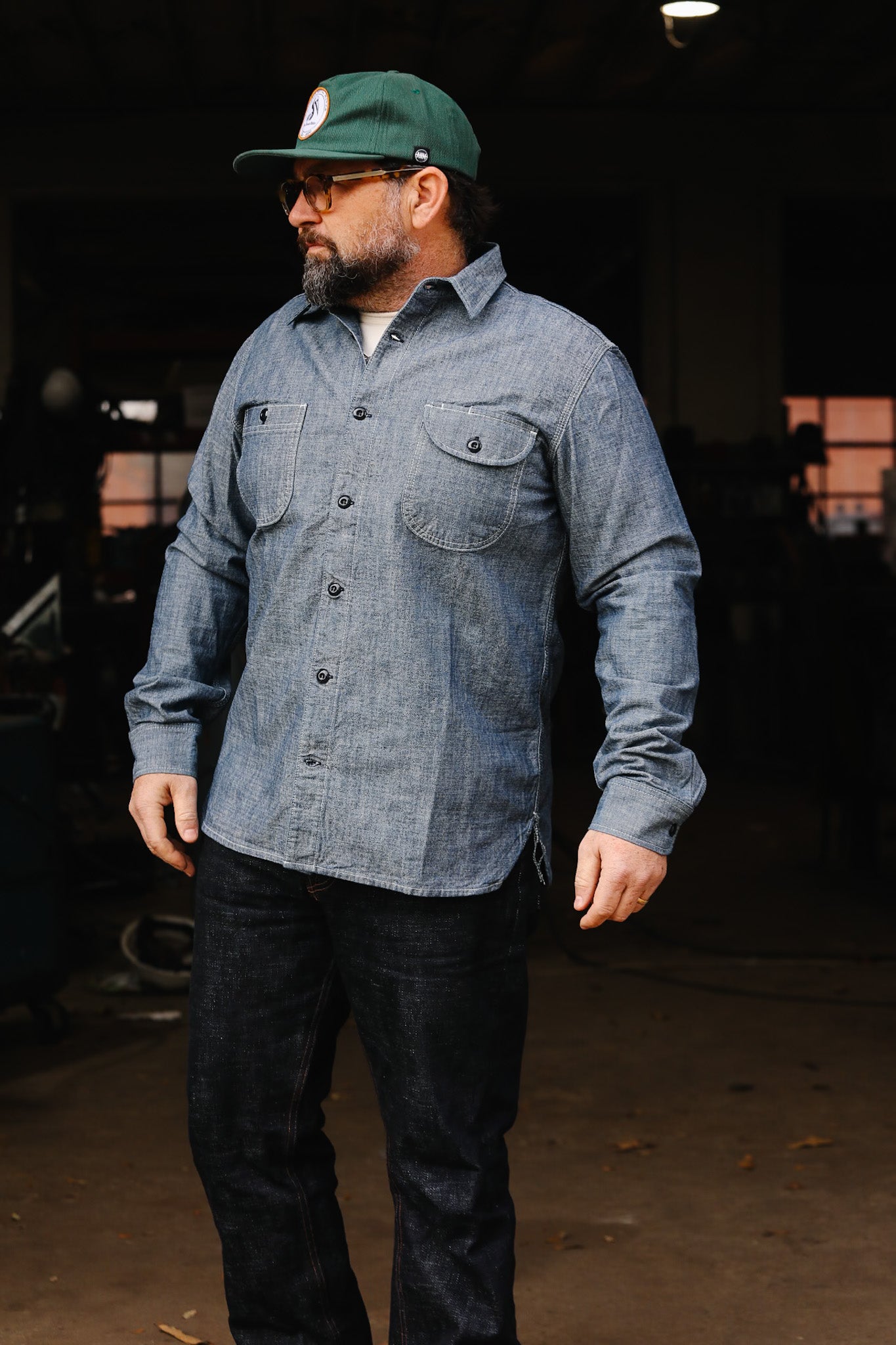 Man wearing denim work shirt and jeans with green cap and glasses in workshop setting
