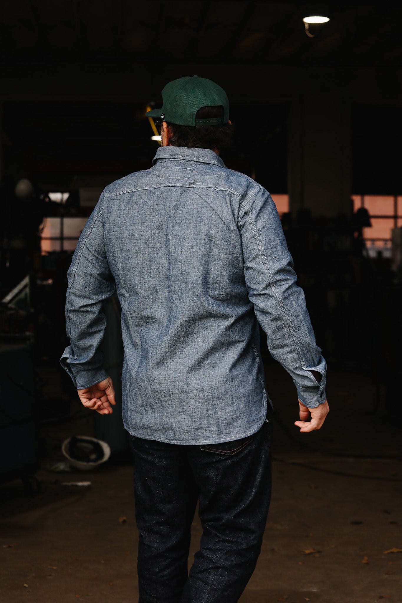 Man wearing denim shirt and green cap standing in industrial workshop