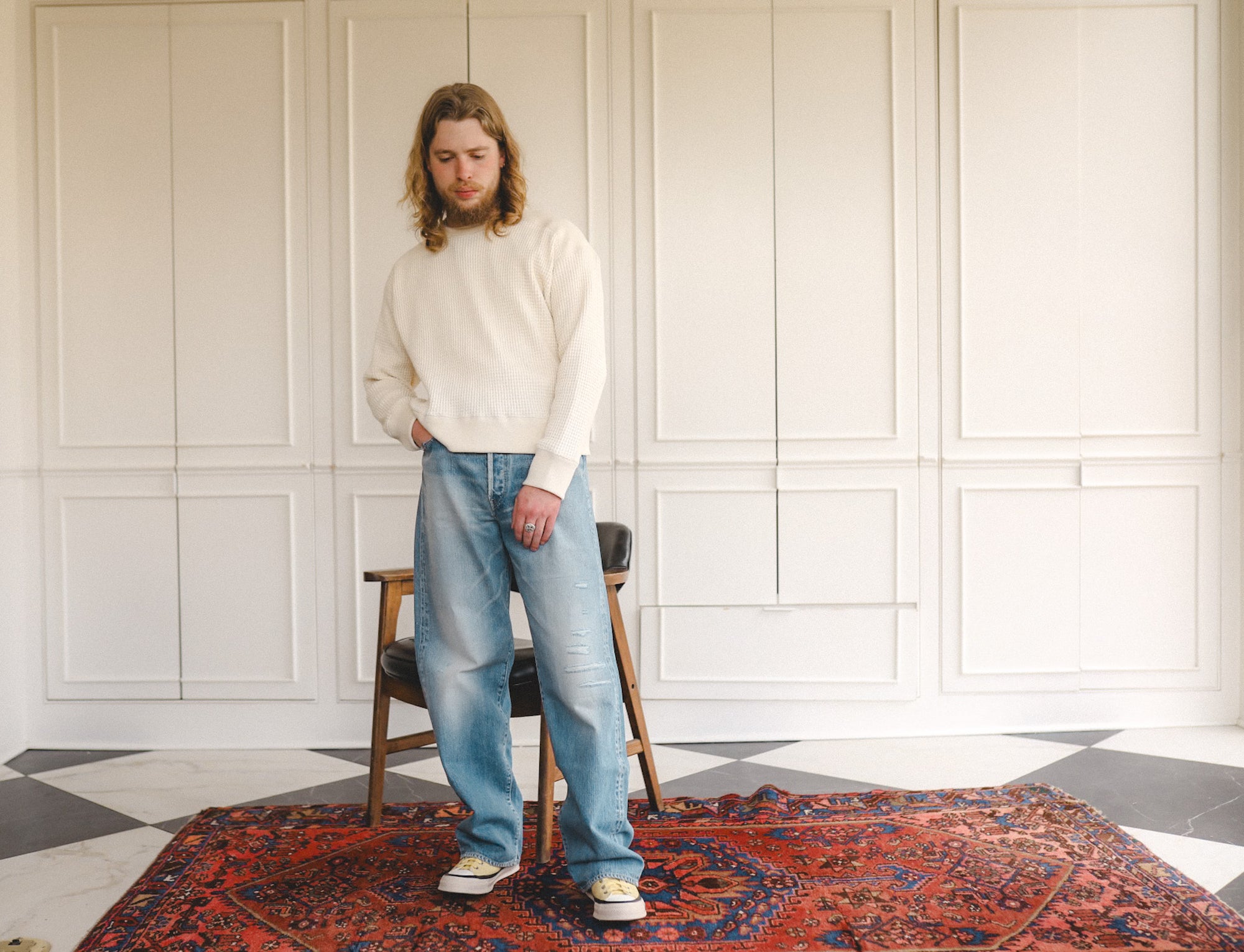 Man wearing a white sweater and blue jeans standing in a room with a patterned rug and wooden chair.