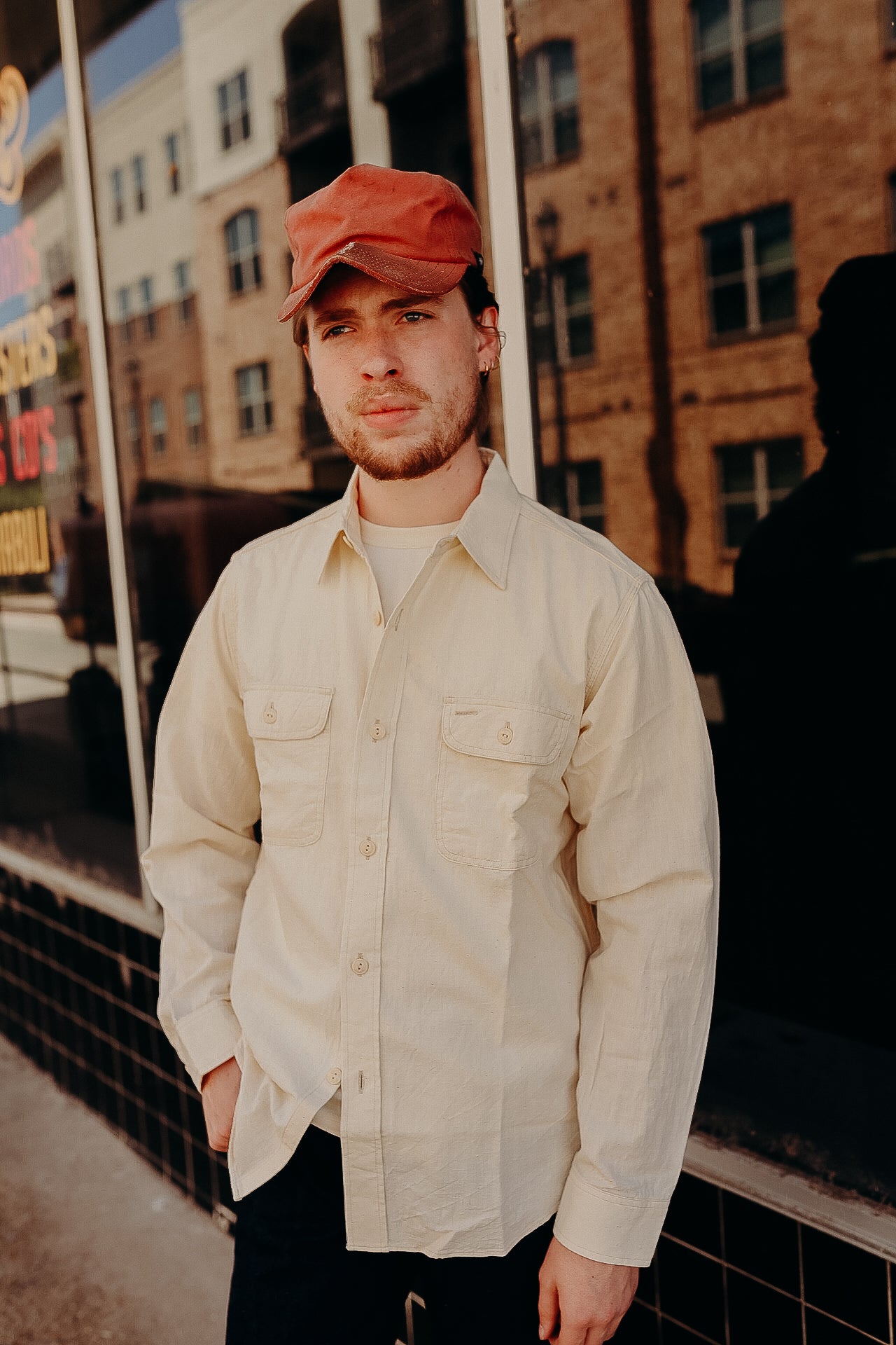 Man wearing beige work shirt and red cap standing outside brick building reflection