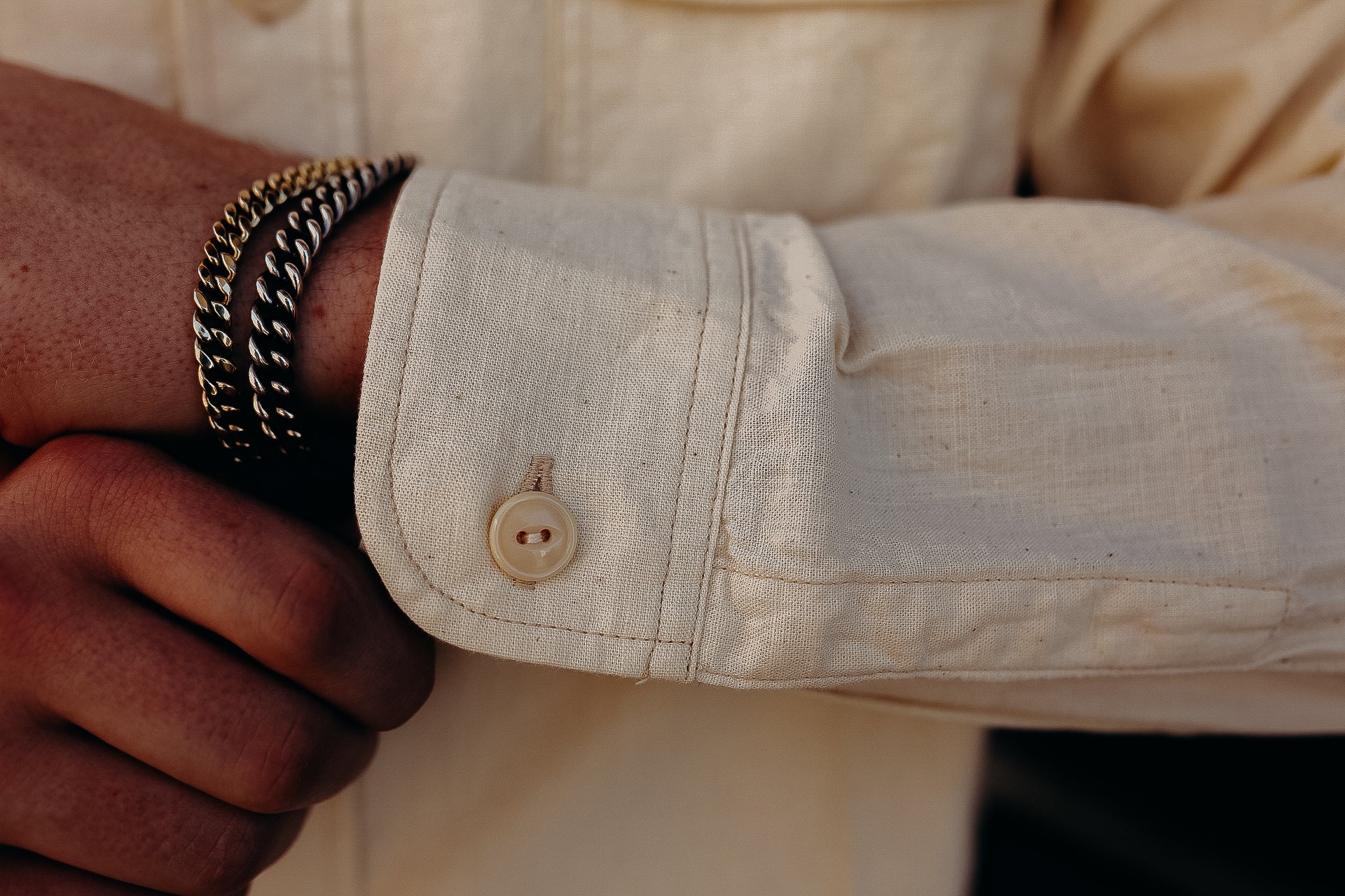 Close-up of a wrist wearing two metal chain bracelets and a beige buttoned shirt cuff