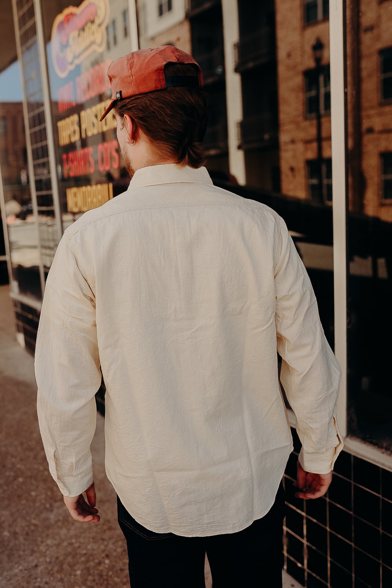 Man wearing beige long-sleeve shirt and red cap walking along urban sidewalk near storefront
