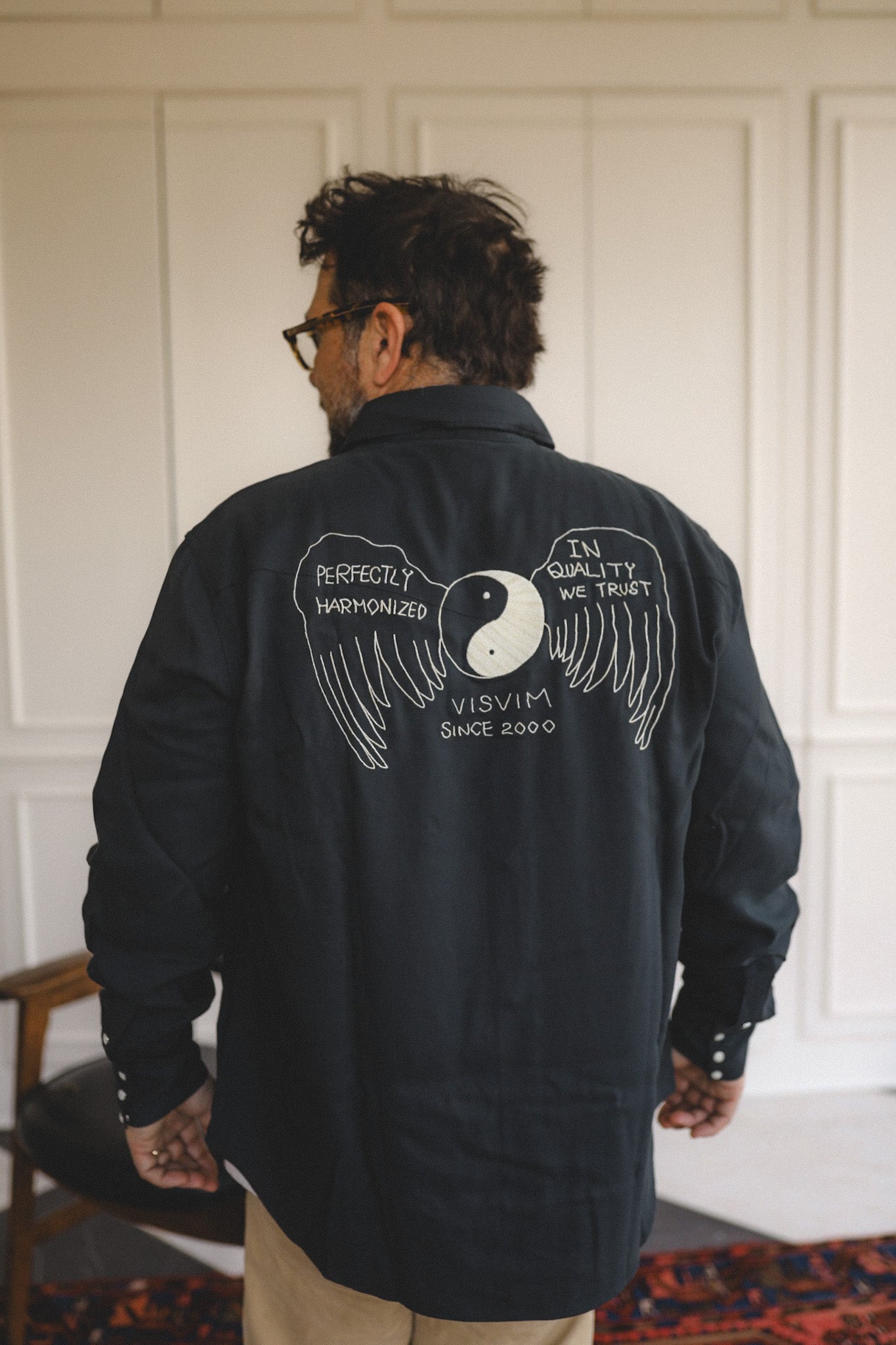 Person wearing a black jacket with white graphics and text on the back, standing against a white paneled wall.
