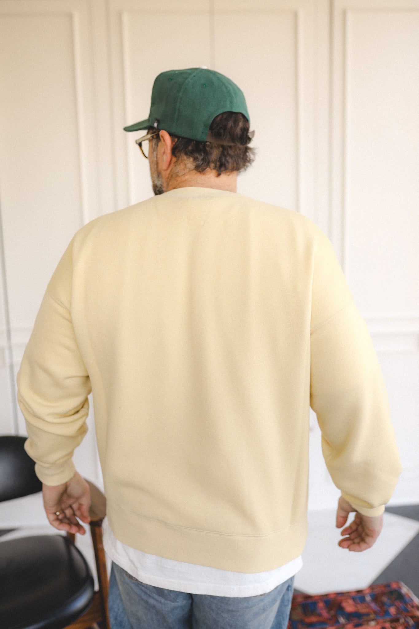 Person wearing a beige sweater and green cap, standing in a room with a chair and rug.