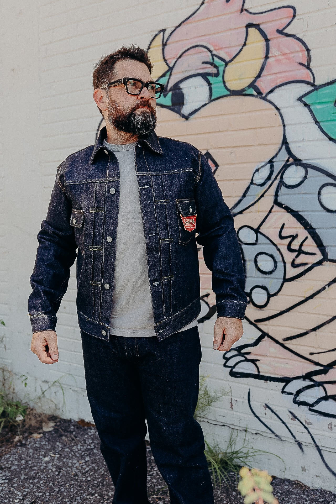 Bearded man wearing dark denim jacket and jeans standing in front of colorful graffiti wall
