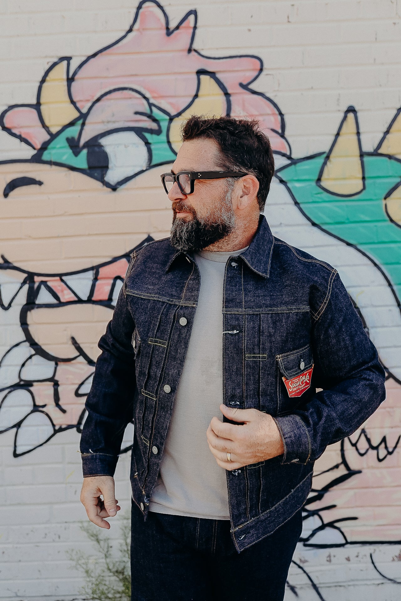 Man with beard and glasses wearing dark denim jacket in front of colorful graffiti wall