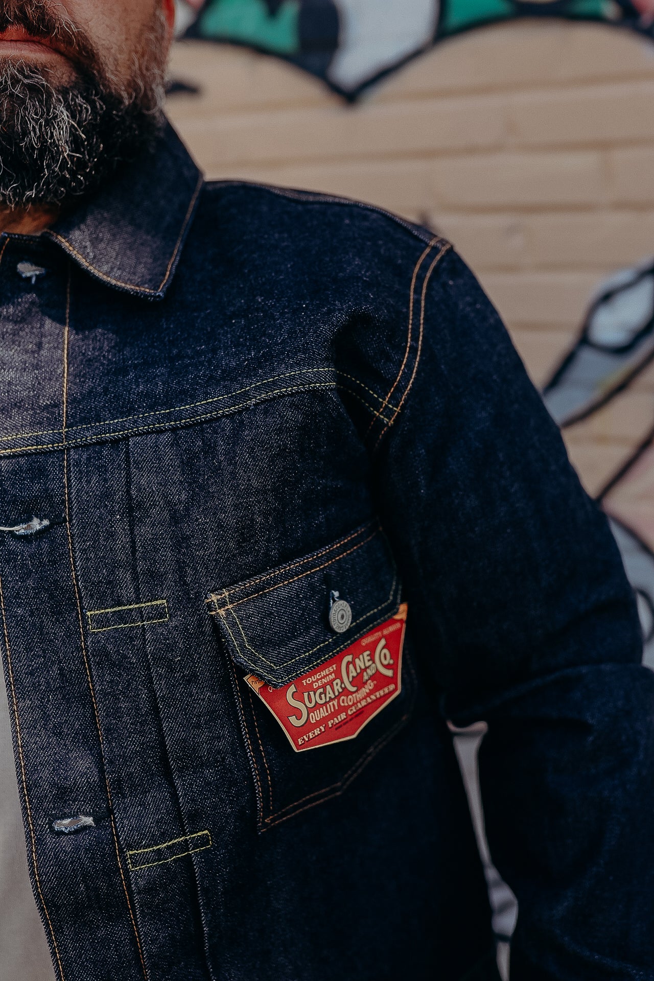 Close-up of man wearing a dark denim jacket with a red Sugar Cane Co label on the chest pocket
