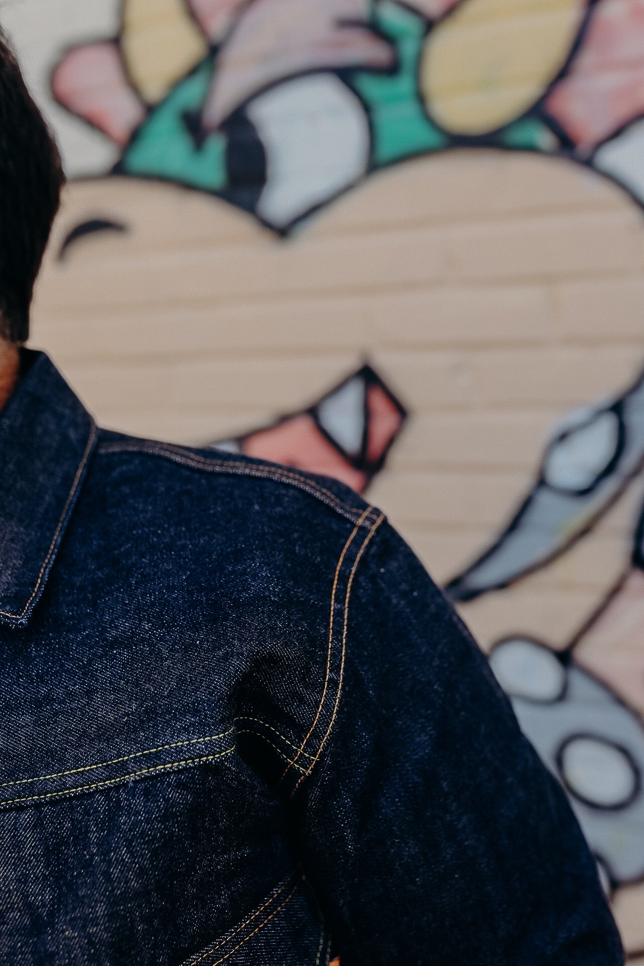 Close-up of denim jacket shoulder with contrast stitching against colorful graffiti wall backdrop