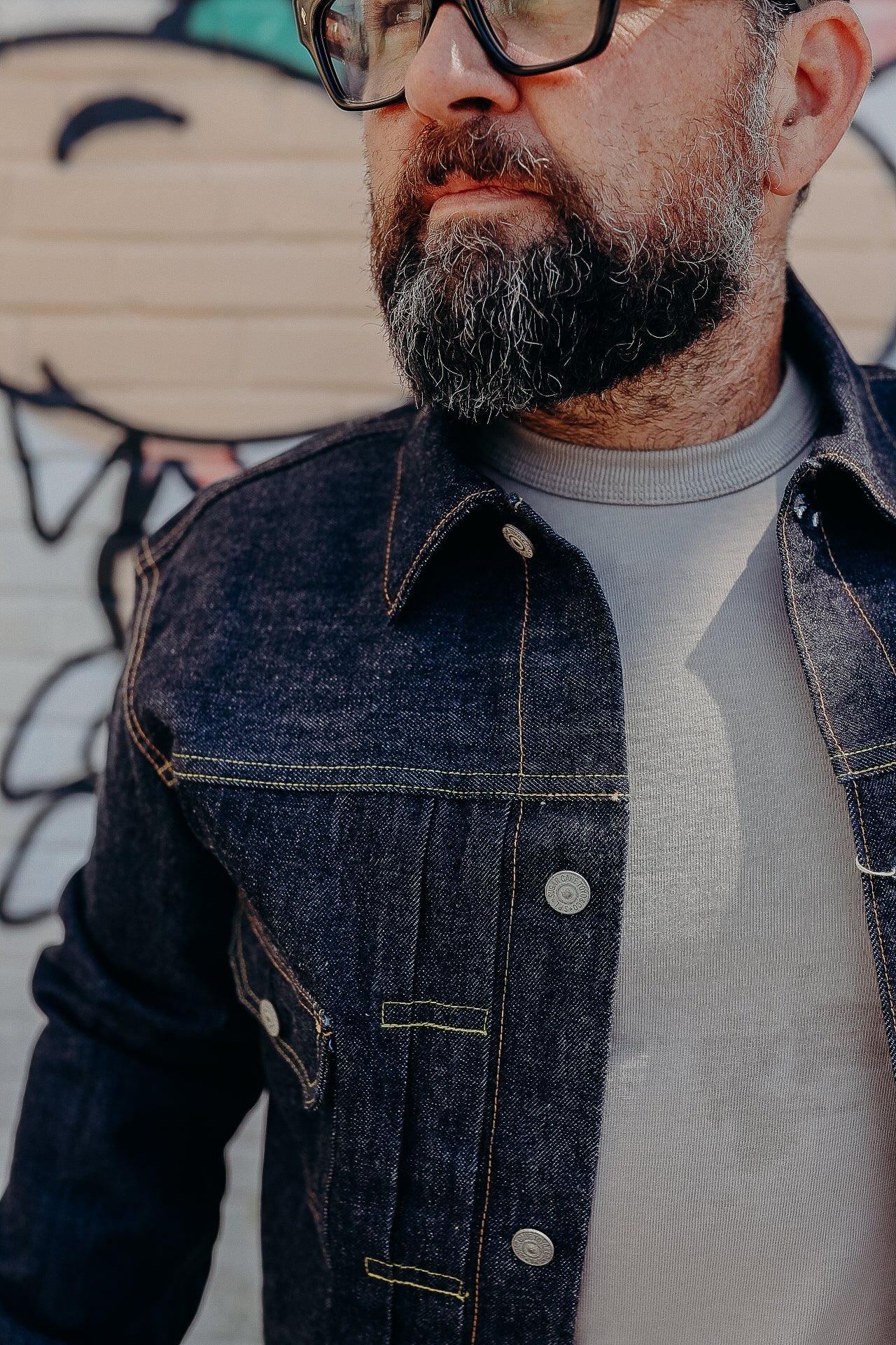 Man with beard wearing black framed glasses and dark denim jacket over gray shirt outdoors