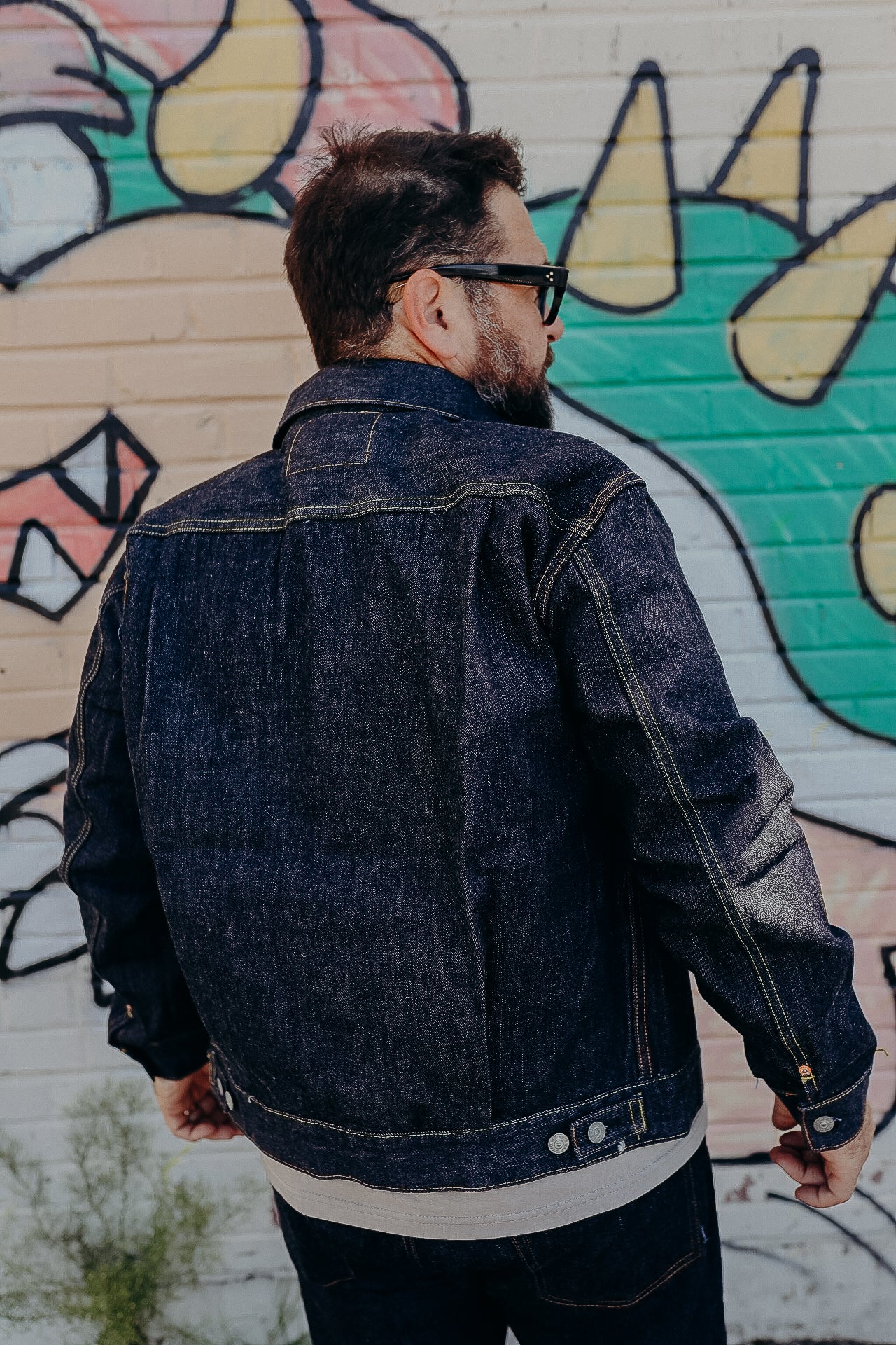 Man with beard and sunglasses wearing dark denim jacket in front of colorful graffiti wall