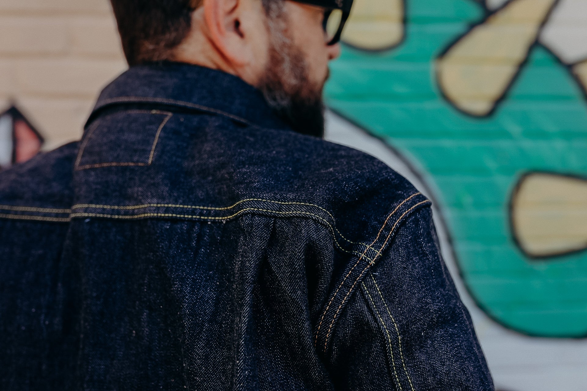 Close-up of a man wearing a dark denim jacket with contrast stitching against a colorful graffiti wall