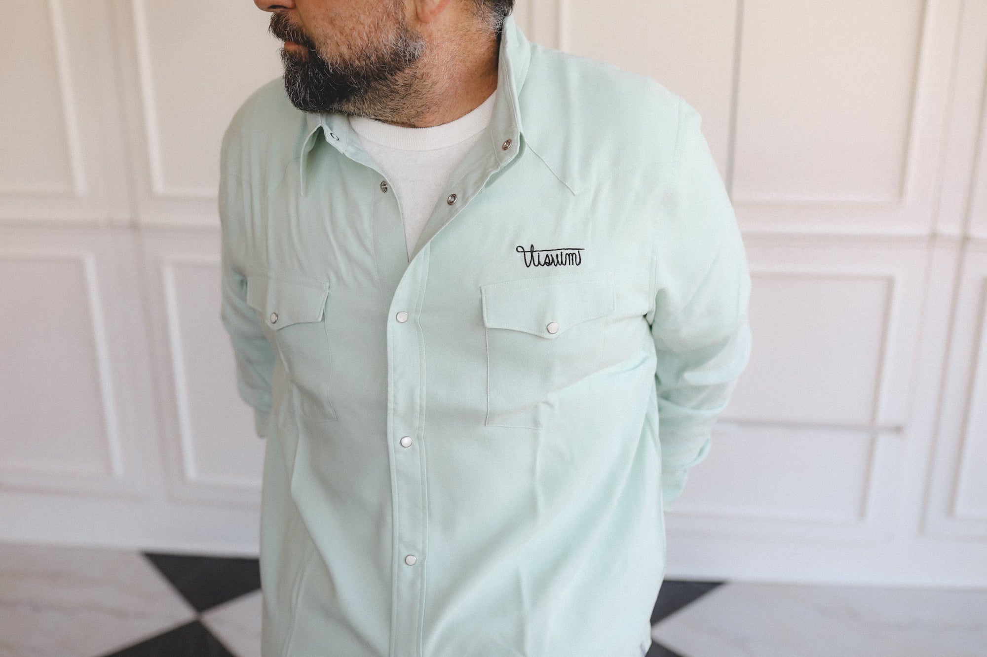 Man wearing a light green button-up shirt with a brand logo against a white paneled wall.
