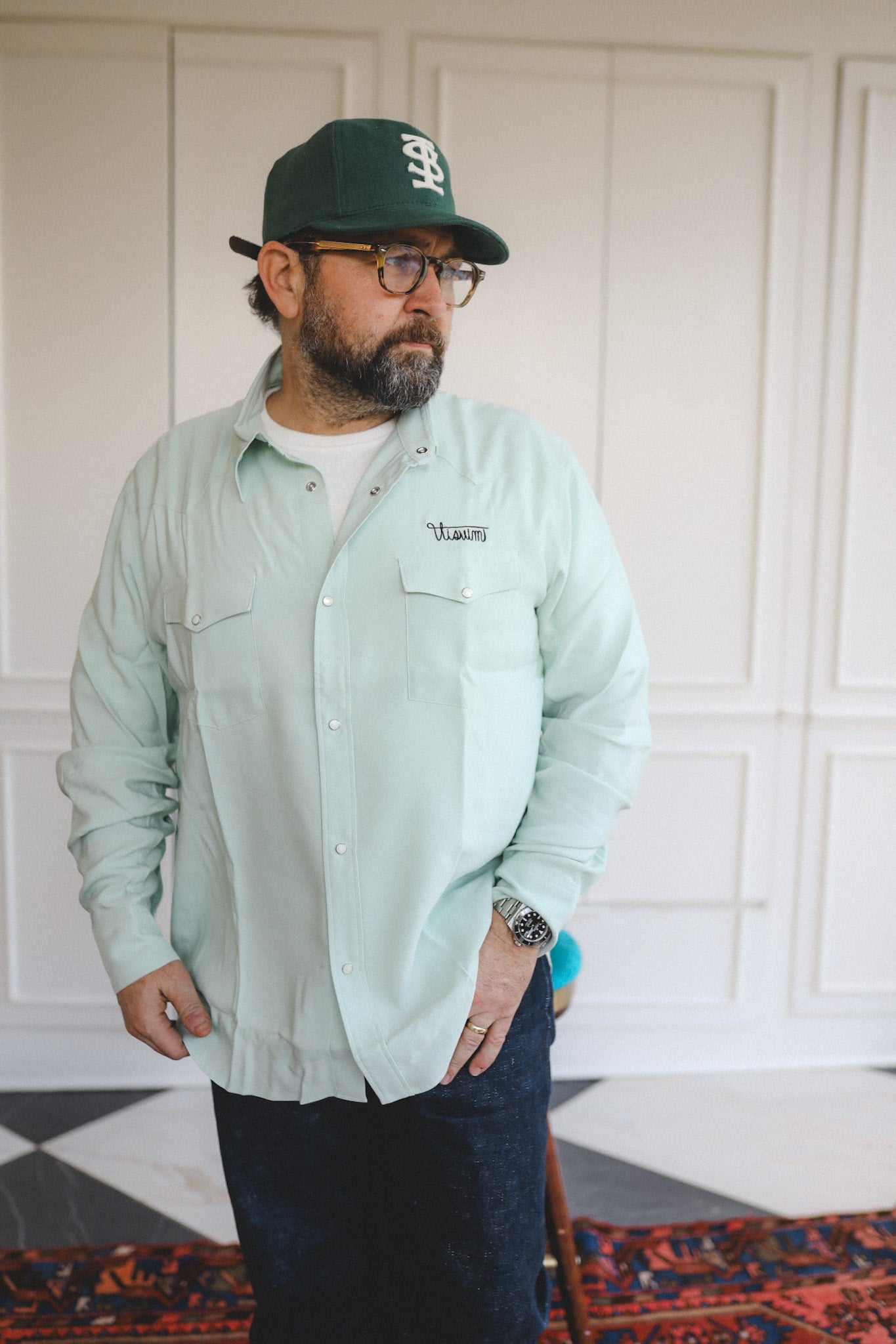 Man wearing a light green shirt, dark cap, and glasses indoors.