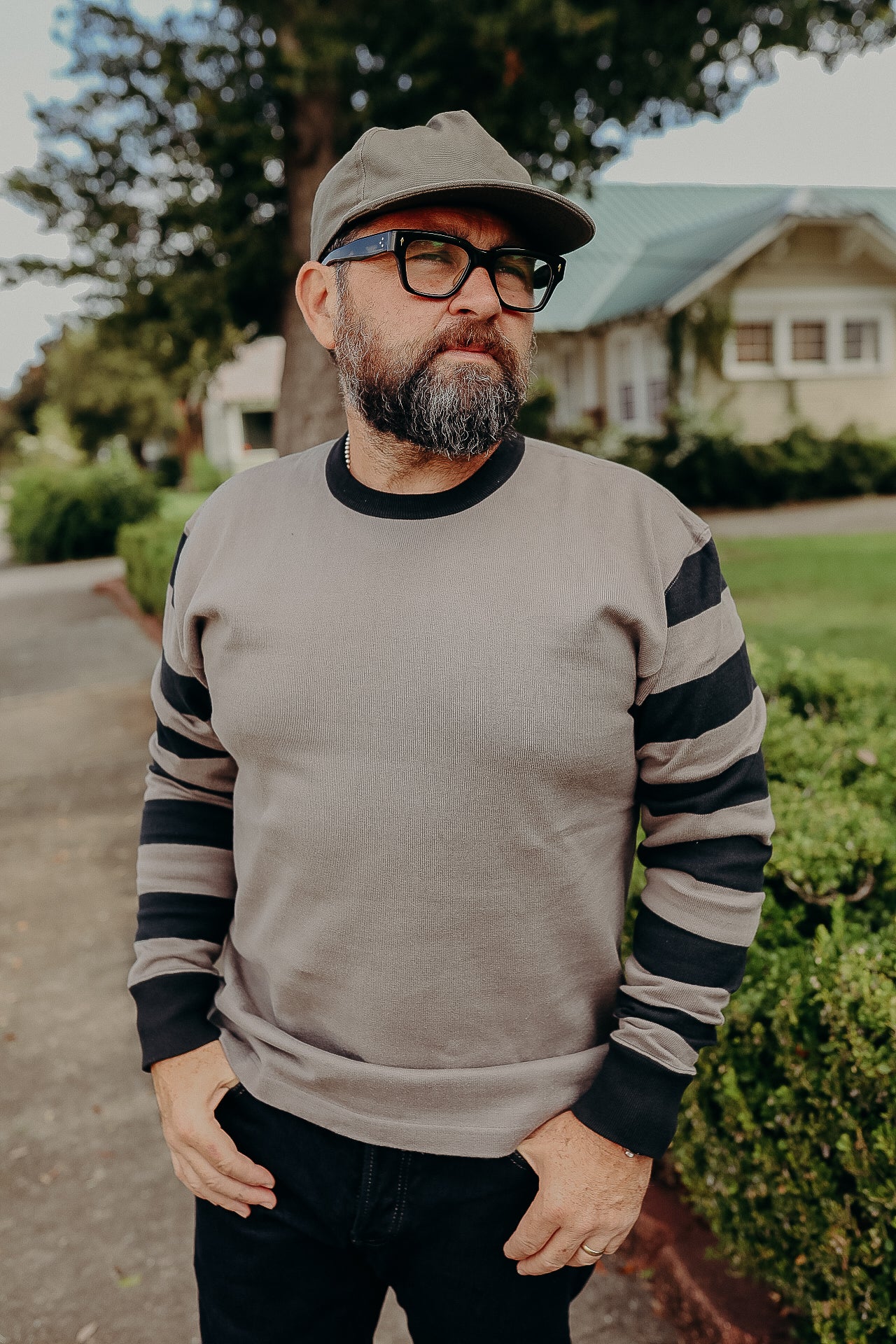 Bearded man wearing glasses, a gray cap, and a gray sweater with black stripes on sleeves outdoors