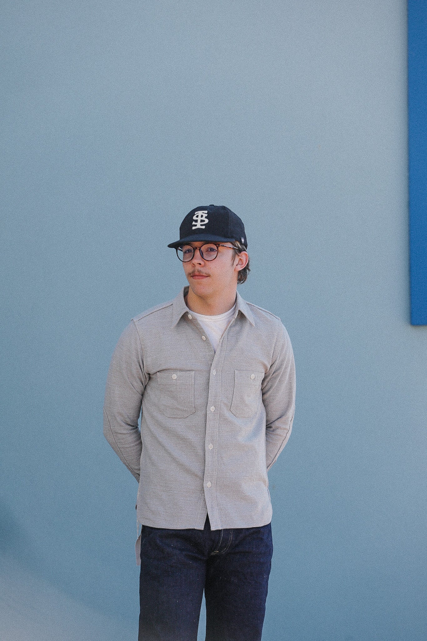 Person wearing a cap and glasses, standing against a light blue wall.