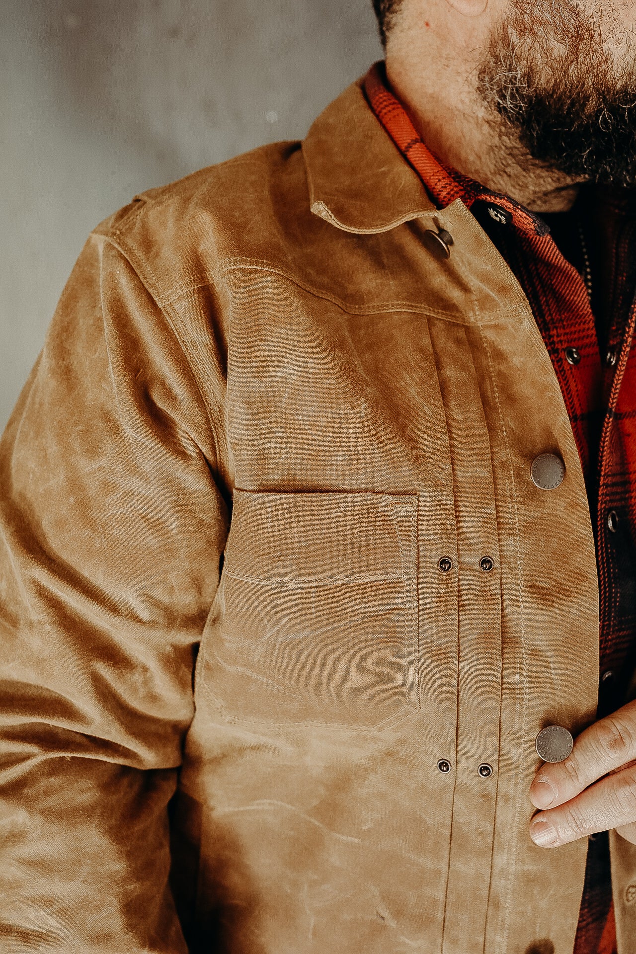 Close-up of man wearing rugged brown waxed canvas work jacket over red plaid shirt, fastening metal snap button
