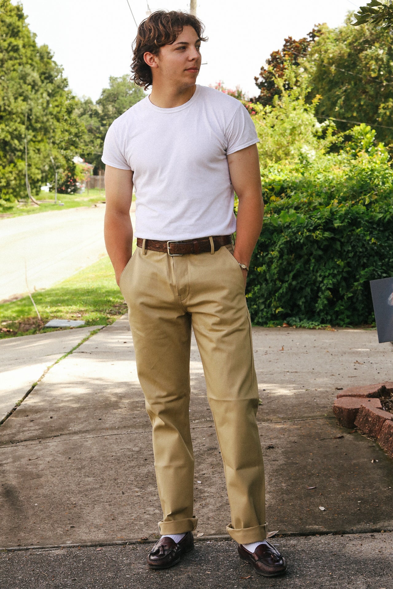 Man wearing white t-shirt, khaki pants with brown belt, and brown loafers standing outdoors on sidewalk