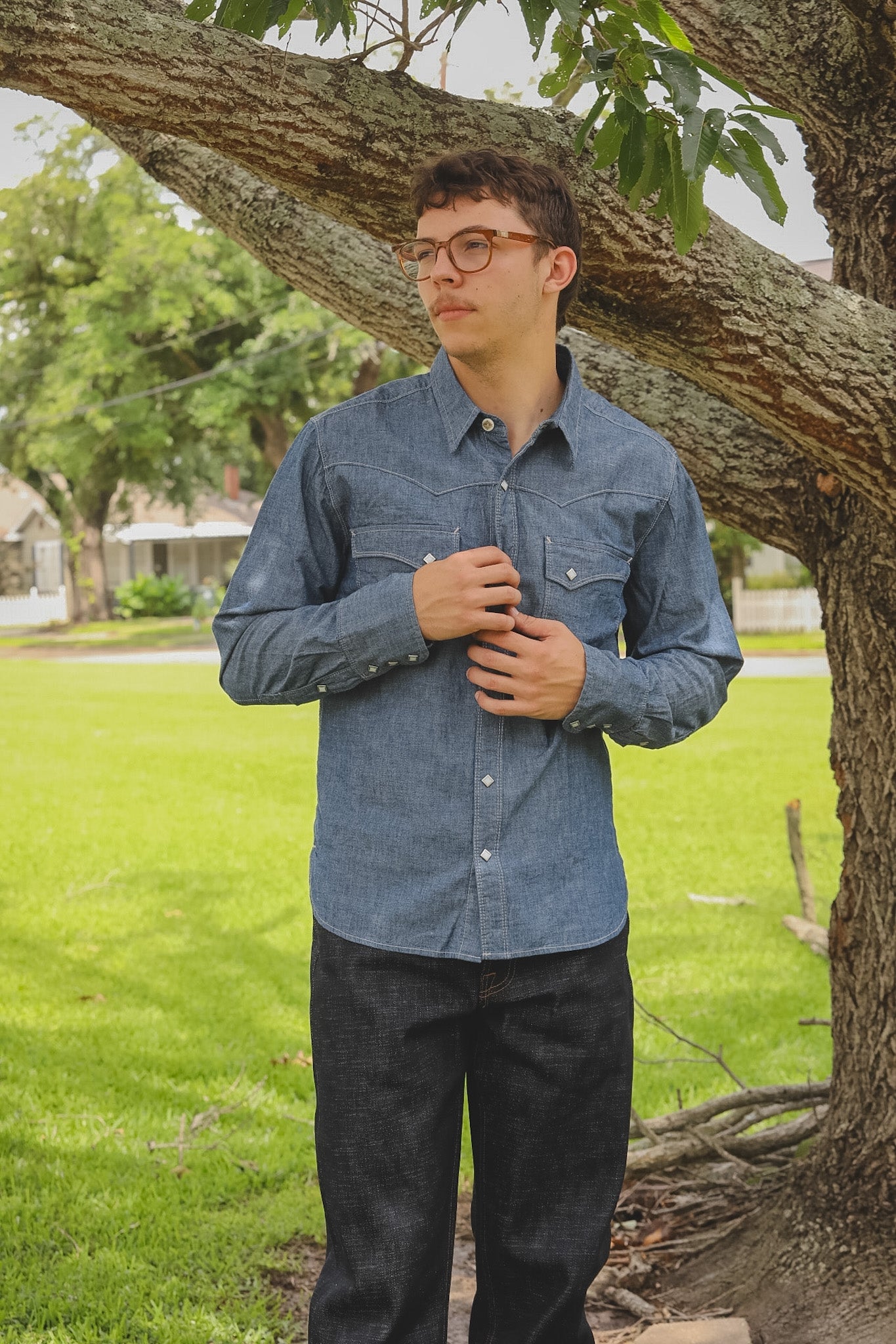 Person wearing a blue denim shirt standing under a tree in a suburban setting
