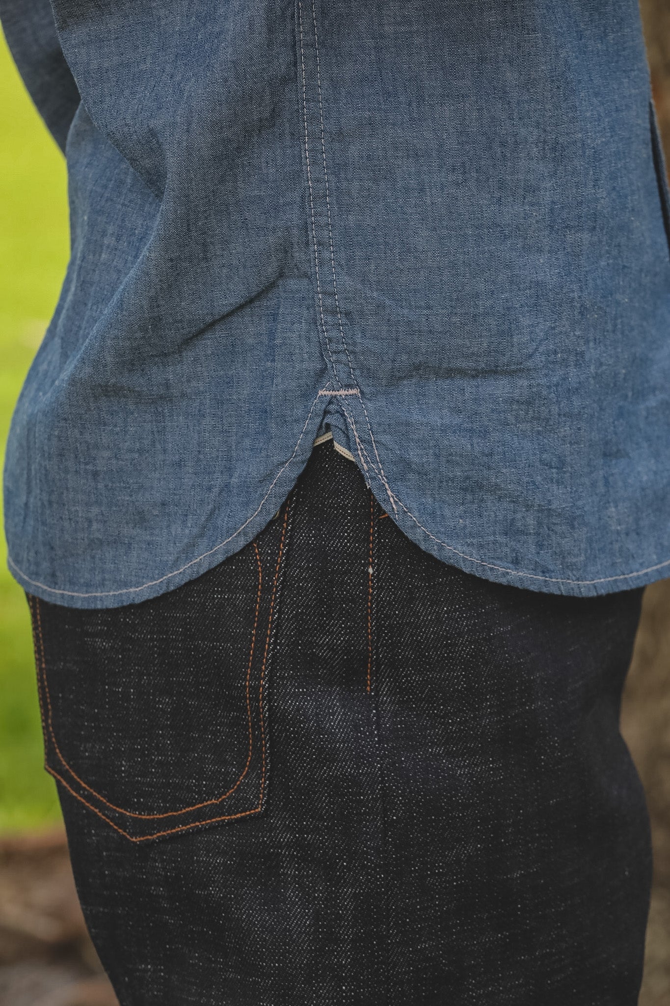Close-up of a person wearing a blue denim shirt and dark jeans with a blurred natural background.